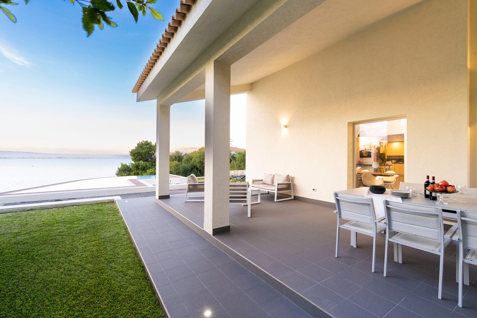 Modern outdoor patio with a dining area overlooking a serene waterfront, featuring stylish furniture, a lush green lawn, and a glimpse of a swimming pool in the background. Ideal for relaxation and entertaining. | Only on SopranoVillas