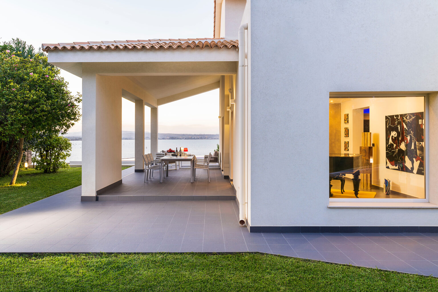 Modern waterfront home exterior showcasing a spacious patio with dining area, lush green lawn, and large windows revealing contemporary interior design, ideal for outdoor living and entertaining. | Only on SopranoVillas