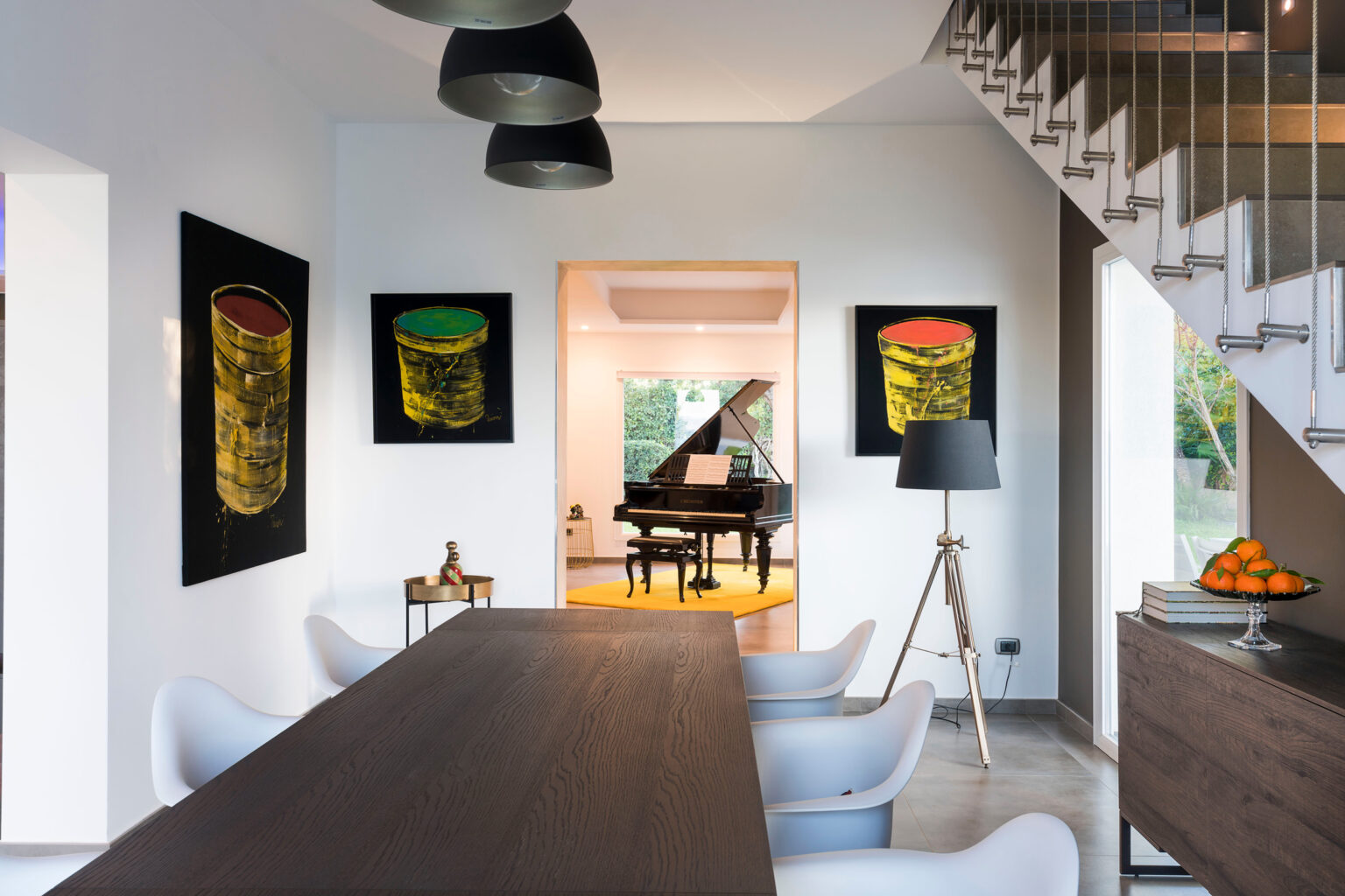 Modern dining area featuring a long wooden table, stylish white chairs, and contemporary artwork on the walls. A grand piano is visible in the background, complemented by a well-lit space with a warm ambiance. | Only on SopranoVillas