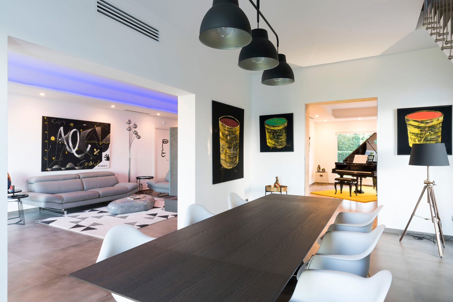Modern dining area featuring a sleek wooden table surrounded by white chairs, with contemporary artwork on the walls and a spacious living room visible in the background. The room is illuminated by stylish pendant lights and has a piano and vibrant decor elements. | Only on SopranoVillas