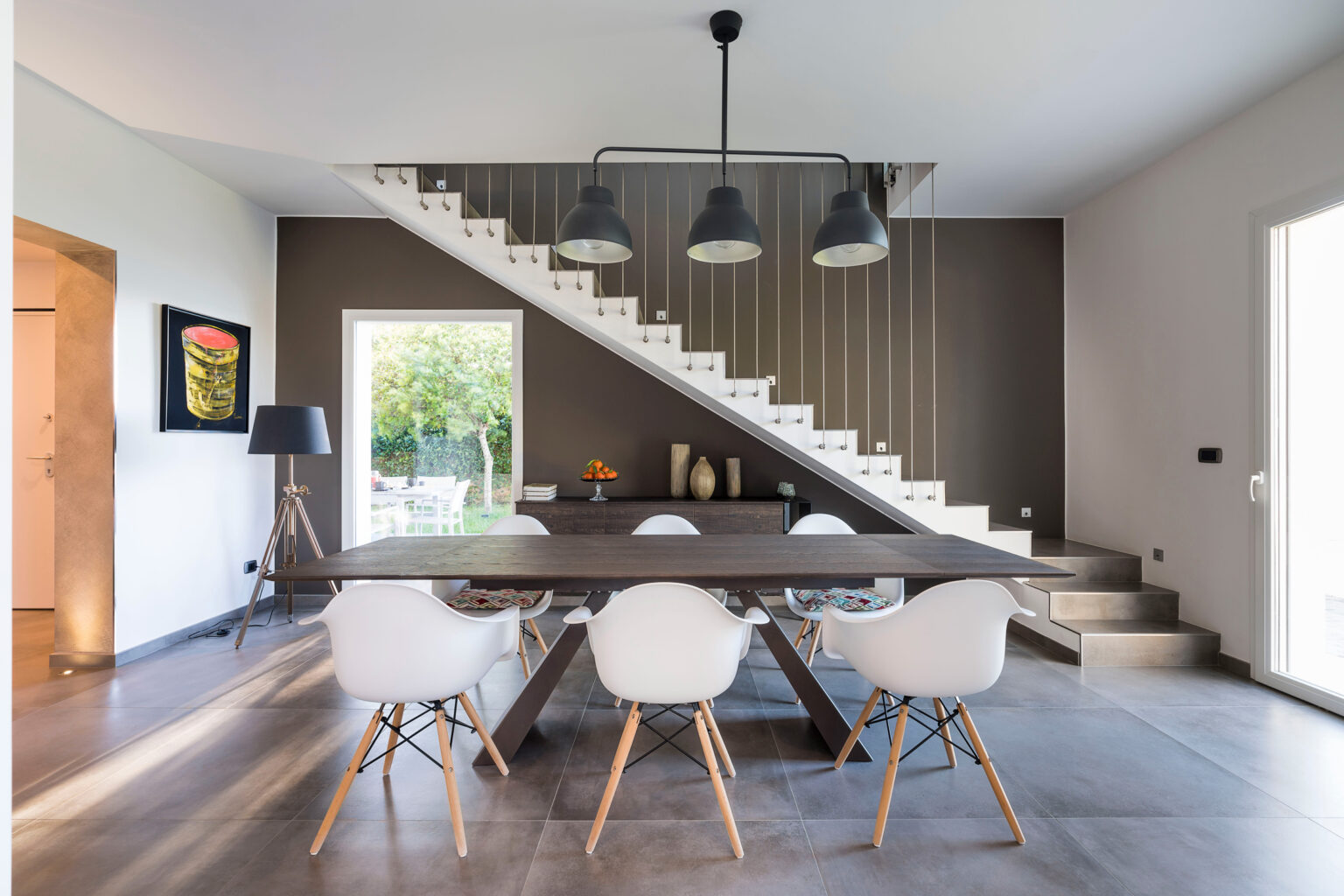 Modern dining room featuring a large wooden table surrounded by white chairs, with a stylish staircase in the background and natural light streaming in through large windows. | Only on SopranoVillas