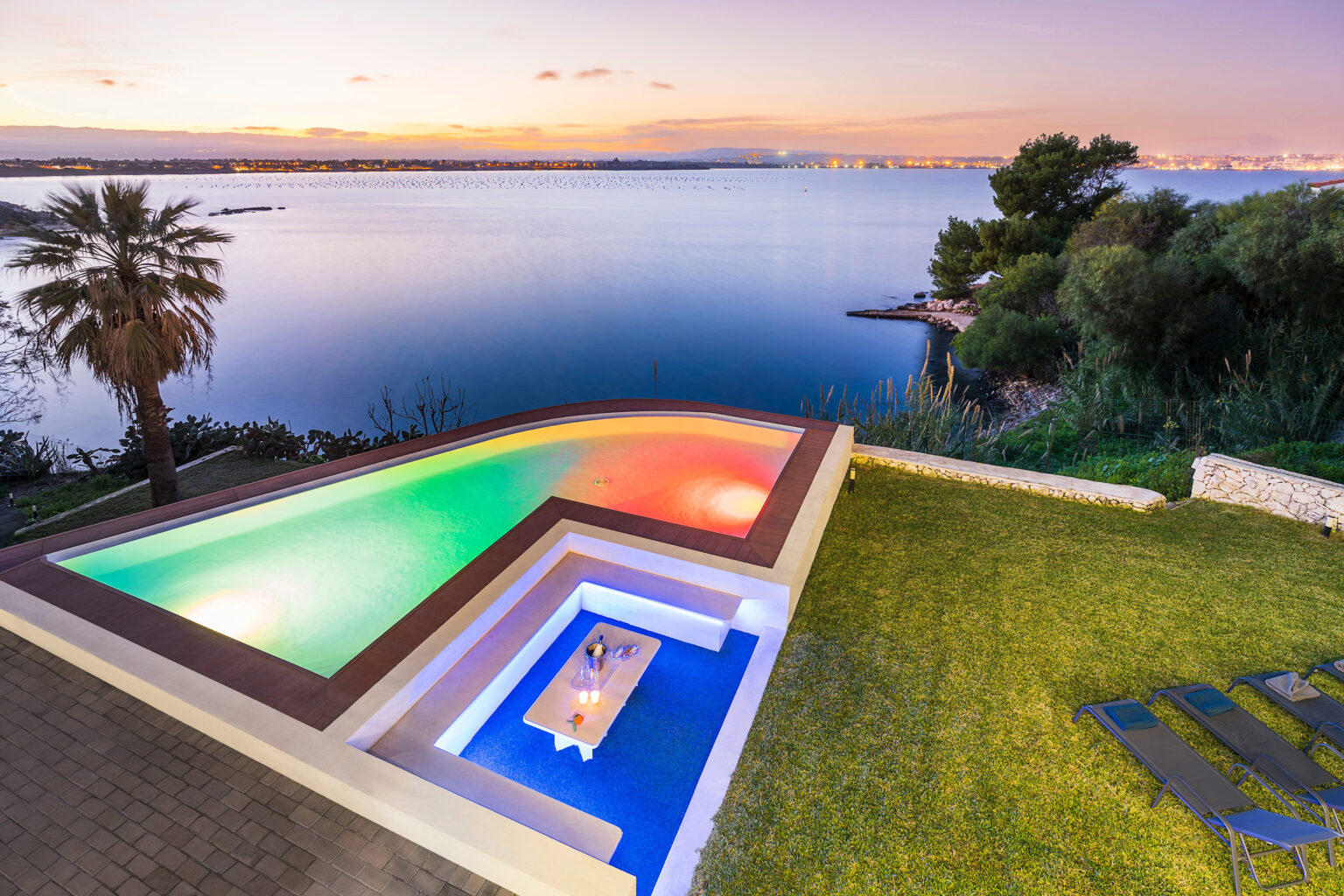 Stunning aerial view of a modern infinity pool with colorful LED lights, surrounded by lush greenery and overlooking a serene lake at sunset. | Only on SopranoVillas