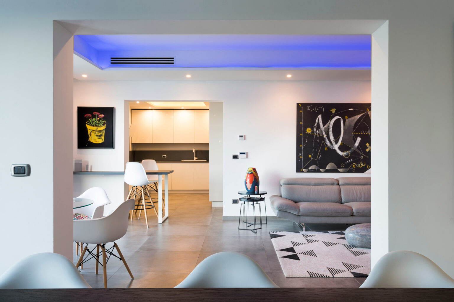 Modern open-concept living space featuring a stylish kitchen with white cabinetry, a cozy gray sofa in the living area, and colorful artwork on the walls. Soft blue lighting enhances the contemporary design, while sleek furniture adds to the inviting atmosphere. | Only on SopranoVillas