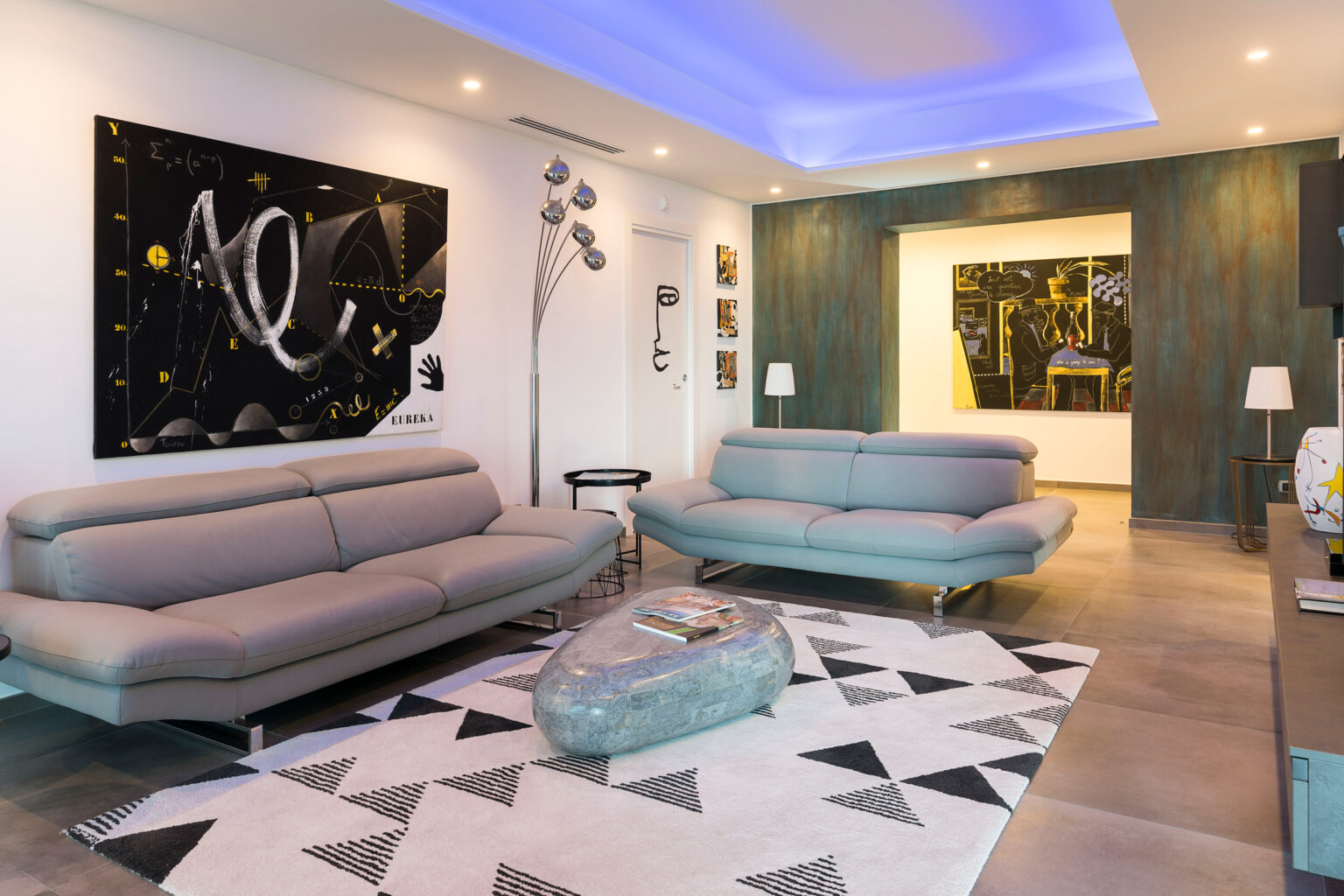 Modern living room featuring two gray sofas, a geometric patterned rug, contemporary artwork on the walls, and ambient lighting. The space is designed with a combination of sleek furniture and artistic decor elements. | Only on SopranoVillas