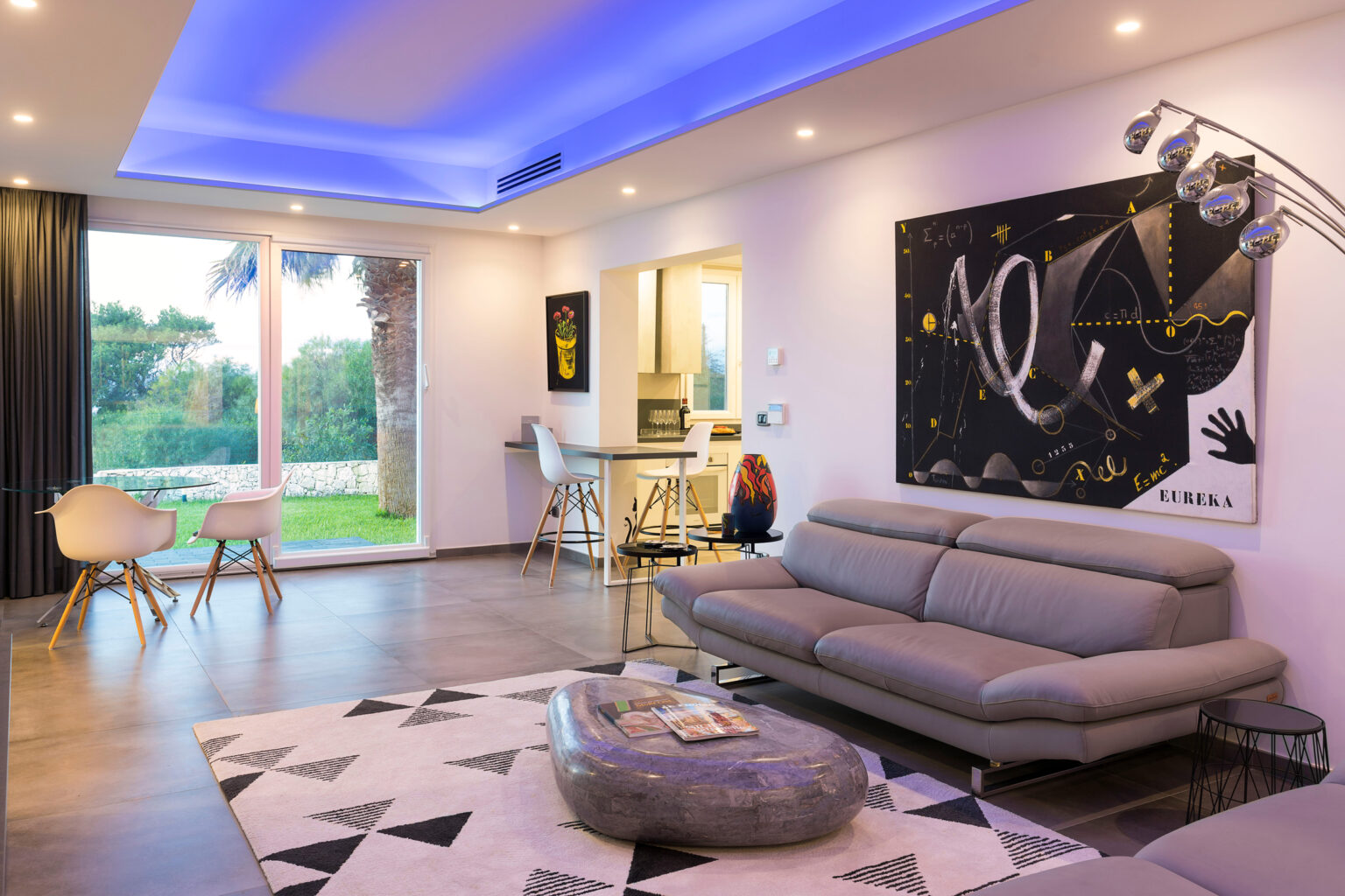 Modern living room featuring a gray sofa, minimalist decor, and artistic wall art. Natural light floods the space through large windows, highlighting a stylish dining area and contemporary furnishings. | Only on SopranoVillas
