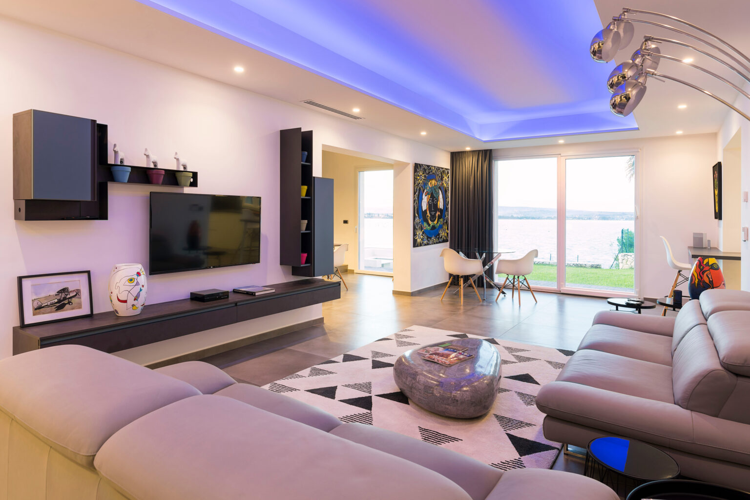 Modern living room with sleek furniture, a large TV, and stylish decor. Features a contemporary sofa, geometric rug, and ambient lighting, creating a cozy atmosphere with a view of the outdoors. | Only on SopranoVillas