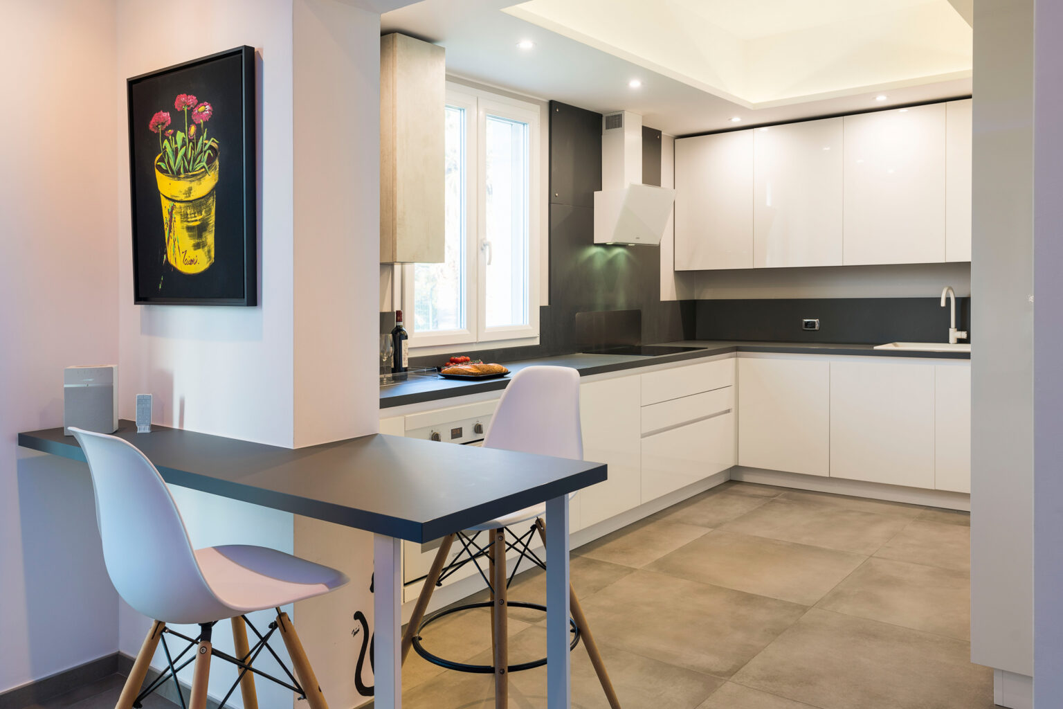 Modern kitchen interior featuring sleek white cabinetry, a black countertop, and a minimalist dining area with stylish bar stools. Natural light floods the space through large windows, complemented by contemporary artwork on the wall. | Only on SopranoVillas