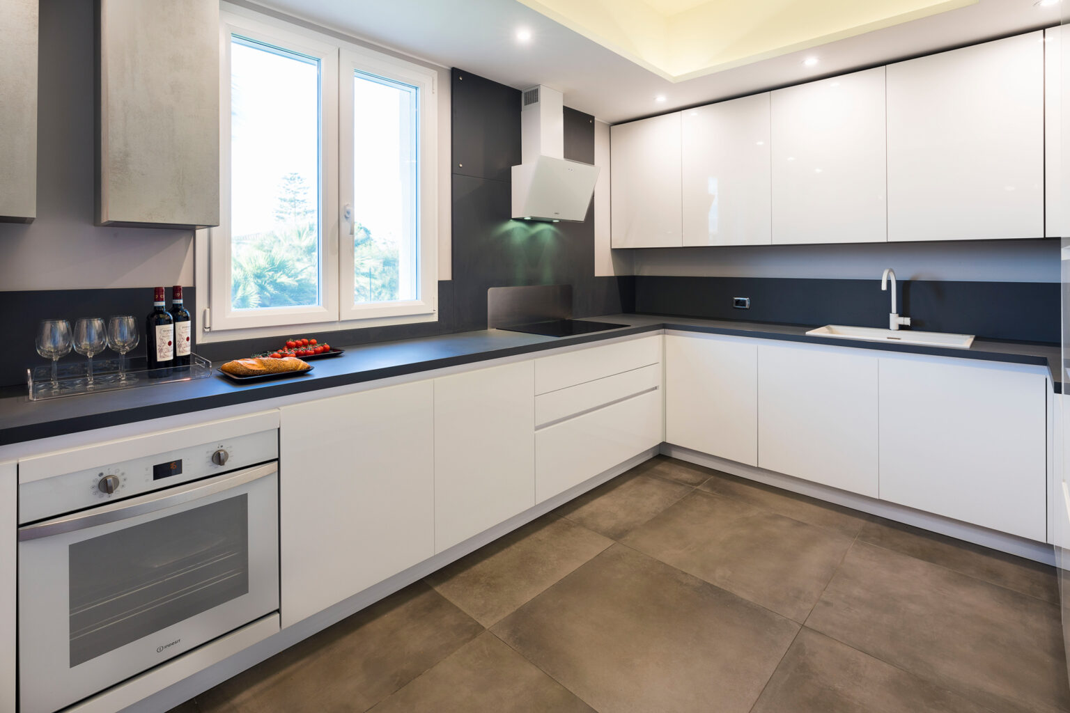 Modern kitchen interior featuring sleek white cabinetry, gray countertops, and stainless steel appliances. Natural light floods the space through a large window, enhancing the contemporary design. A tray with snacks and wine bottles adds a welcoming touch. | Only on SopranoVillas
