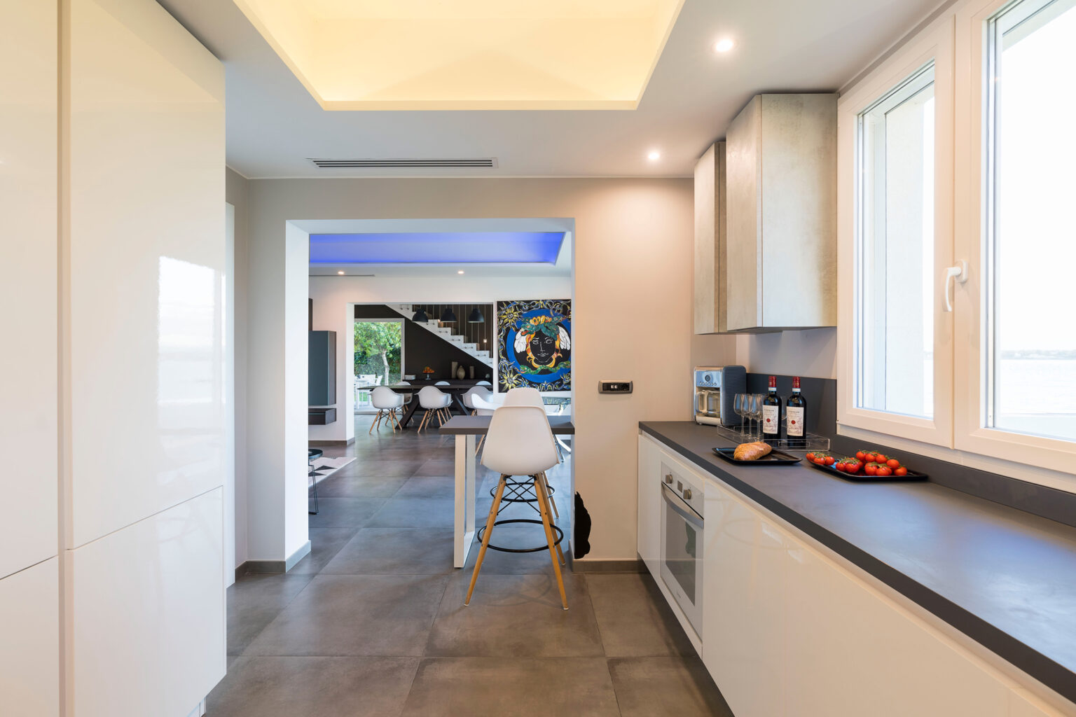 Modern kitchen interior featuring sleek cabinetry, a breakfast bar with a white stool, and large windows allowing natural light. The open layout connects to a stylish living area, showcasing contemporary design elements. | Only on SopranoVillas