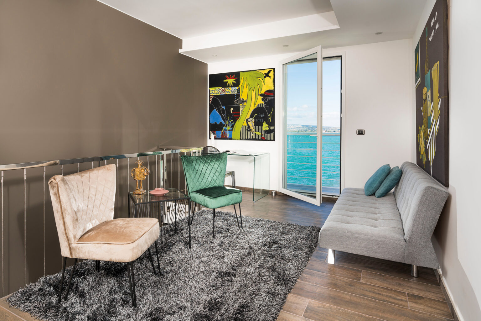 Modern living room with stylish seating including a beige armchair, green armchair, and gray sofa, set against a backdrop of large windows showcasing a scenic water view. The room features contemporary decor, a plush rug, and vibrant artwork on the walls. | Only on SopranoVillas