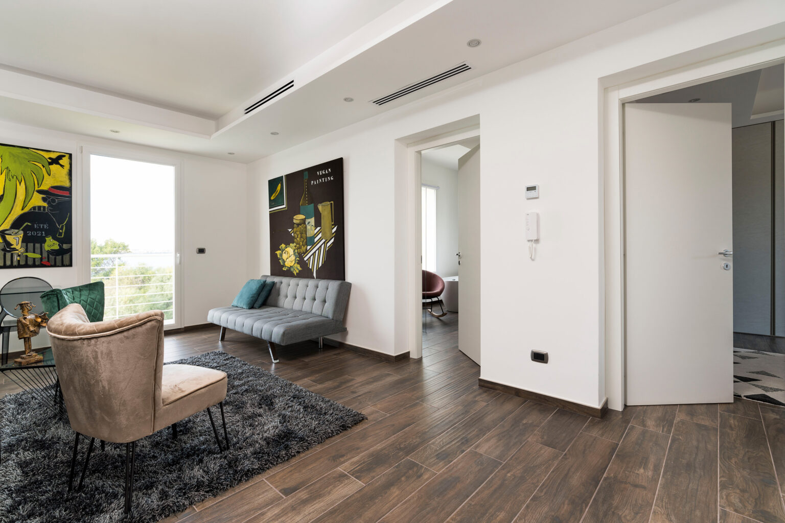 Modern living room interior featuring a grey sofa, beige accent chair, and vibrant artwork on white walls. The space has wooden flooring, a plush area rug, and large windows allowing natural light. A doorway leads to another room, enhancing the open layout. Ideal for contemporary home decor inspiration. | Only on SopranoVillas
