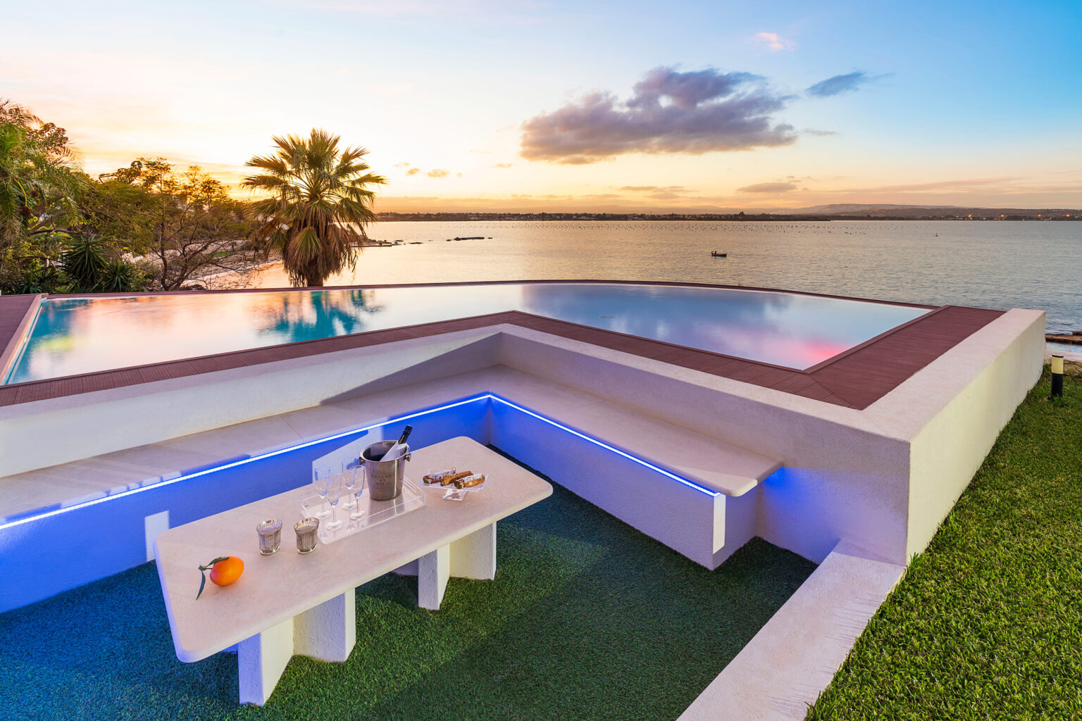 Modern infinity pool overlooking a serene sunset by the water, featuring a stylish outdoor seating area with a table and drinks, surrounded by lush greenery. | Only on SopranoVillas