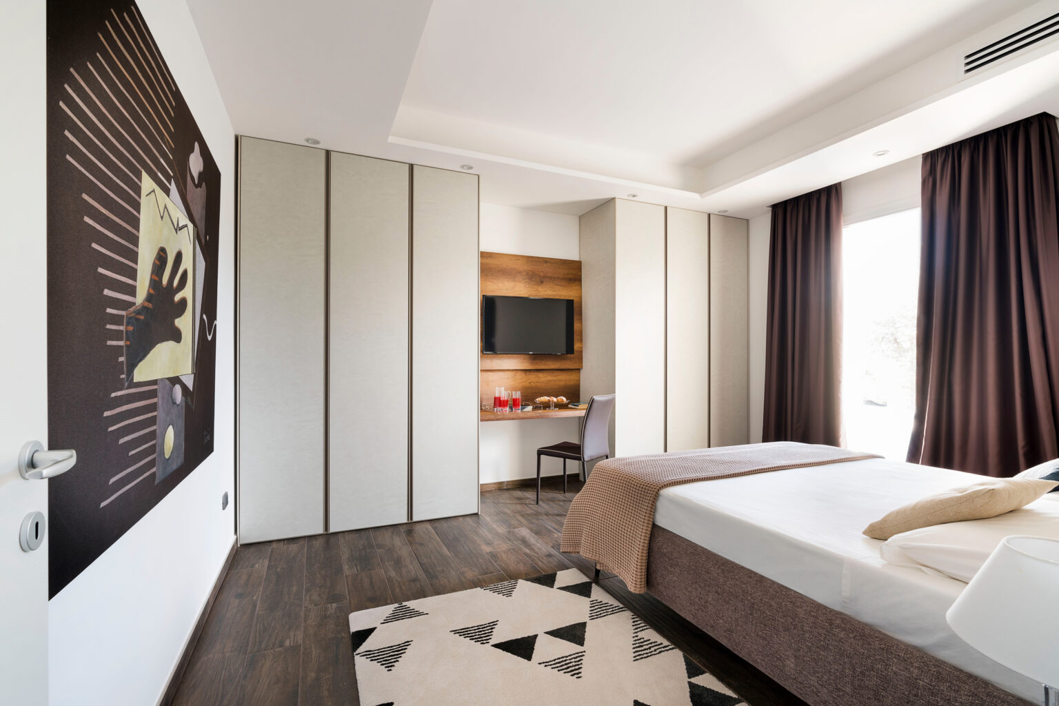 Modern hotel room featuring a minimalist design, hardwood flooring, a large bed with neutral bedding, a stylish area rug, and a wall-mounted TV. The room is illuminated by natural light from large windows covered with dark curtains, complemented by contemporary artwork on the wall. | Only on SopranoVillas