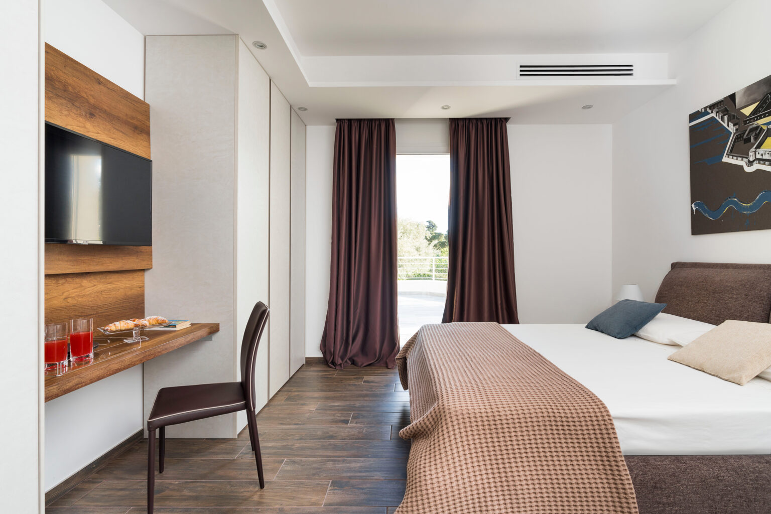 Modern hotel room featuring a queen bed with decorative pillows, a wooden accent wall with a flat-screen TV, and a small workspace with a chair. The room has large windows with brown curtains, allowing natural light, and offers a view of the outdoors. A side table displays refreshments, including glasses of red drinks and pastries. The flooring is designed with wooden tiles, contributing to a contemporary aesthetic. | Only on SopranoVillas