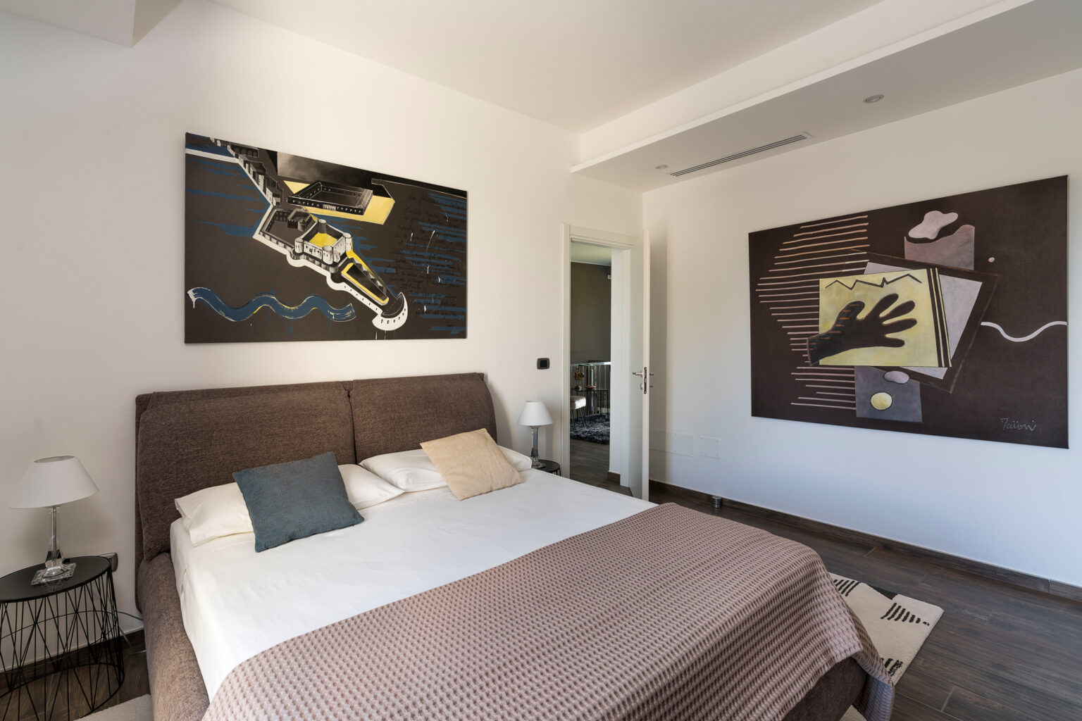 Modern bedroom featuring a stylish bed with decorative pillows, contemporary artwork on the walls, and a minimalist design. The room is bright and spacious, showcasing a blend of comfort and artistic flair. | Only on SopranoVillas