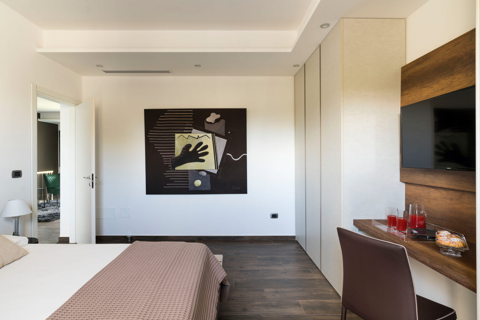 Modern bedroom interior featuring a stylish design with a large abstract painting, wooden accents, and a cozy bed with a textured throw. The room includes a flat-screen TV mounted on the wall, a small workspace with drinks and snacks, and an open door leading to a well-lit area. | Only on SopranoVillas