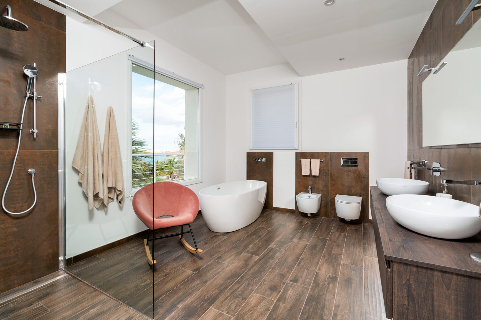 Modern bathroom interior featuring a freestanding bathtub, a glass shower enclosure, dual sinks, and a stylish rocking chair. Natural light streams through a large window, highlighting the wood-like tile flooring and contemporary design elements. | Only on SopranoVillas