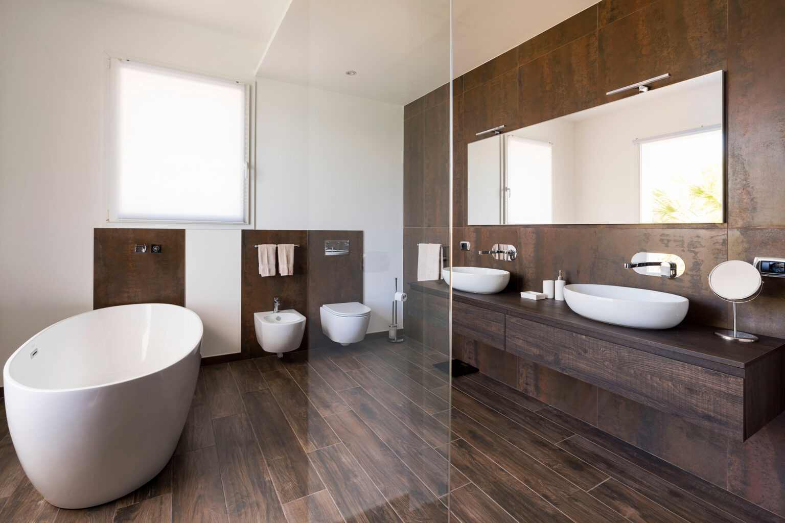Modern bathroom featuring a freestanding bathtub, dual sinks, a large mirror, and sleek wooden cabinetry. Natural light floods the space through a window, enhancing its contemporary design and open layout. | Only on SopranoVillas