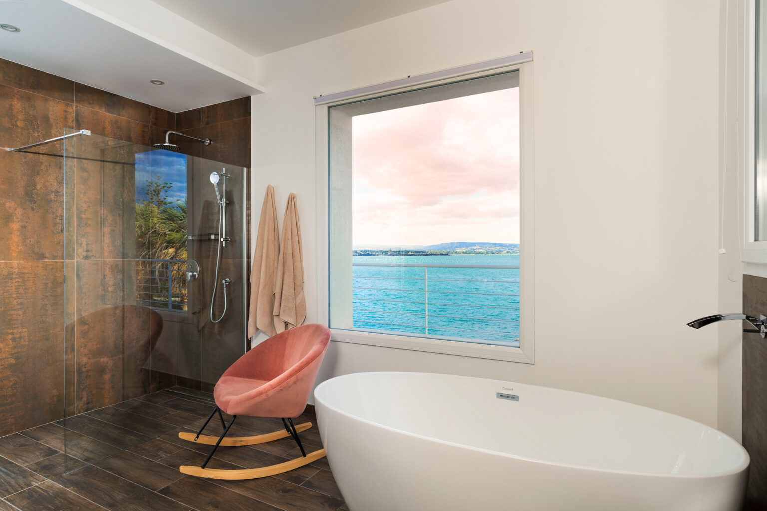 Modern bathroom interior featuring a glass shower, a freestanding white bathtub, and a cozy pink rocking chair. Natural light floods in through a large window overlooking a scenic waterfront view. Neutral tones and wood accents create a serene and stylish atmosphere. | Only on SopranoVillas