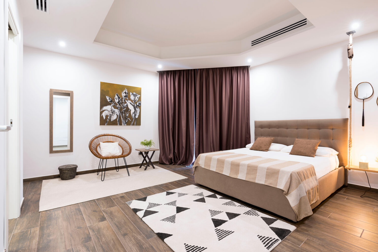 Modern bedroom interior featuring a comfortable bed with neutral bedding, a stylish armchair, a decorative mirror, and elegant curtains, complemented by wooden flooring and contemporary artwork. | Only on SopranoVillas