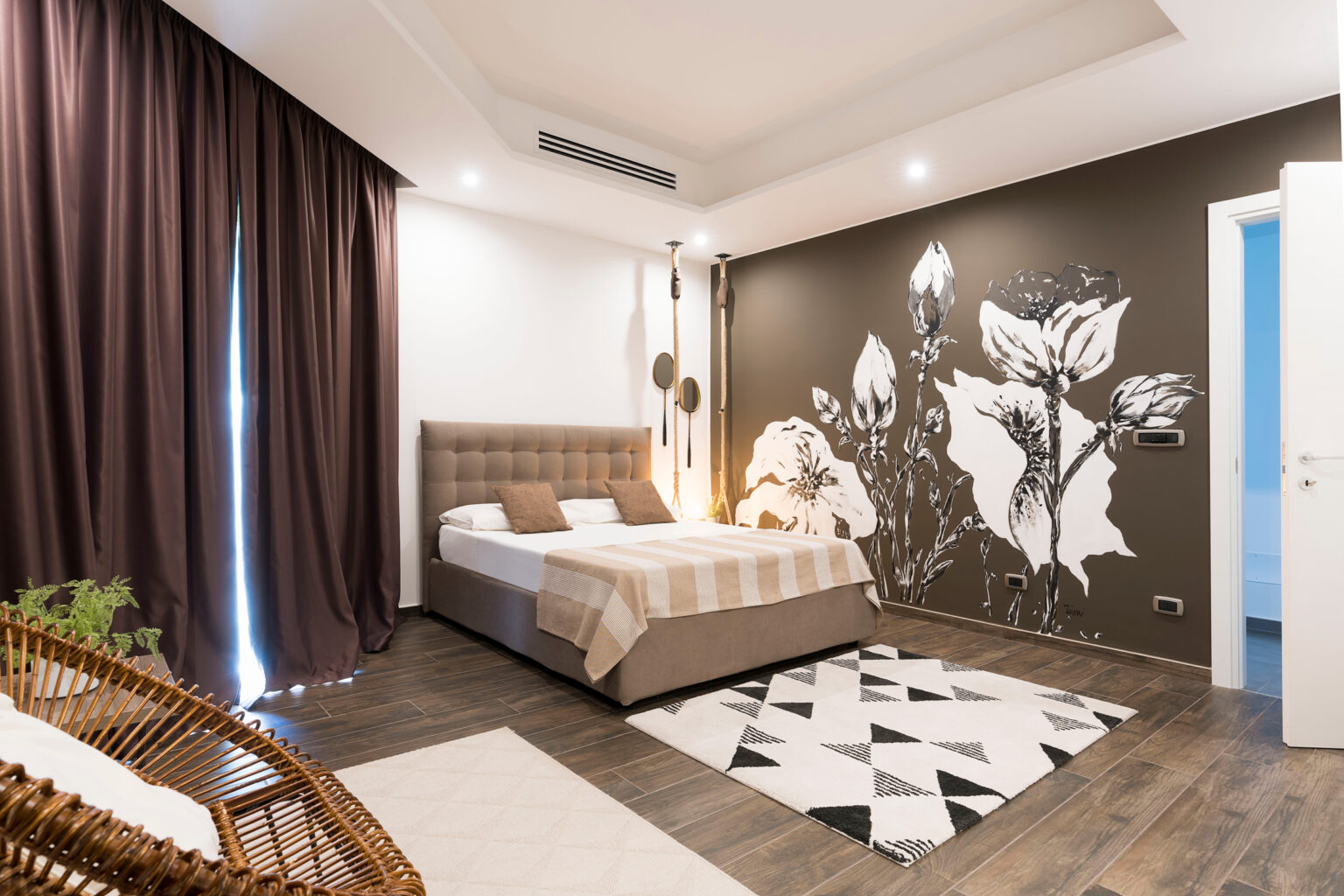 Modern bedroom interior featuring a queen-sized bed with a beige and white striped duvet, dark curtains, and a floral accent wall. The room includes wooden flooring, a geometric patterned rug, and a cozy seating area with a wicker chair, creating a stylish and inviting atmosphere. | Only on SopranoVillas