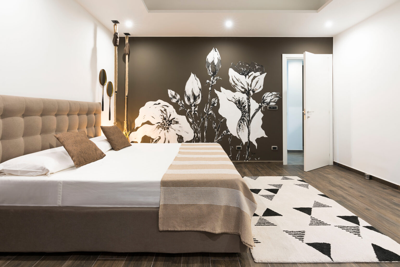 Modern hotel room featuring a stylish design with a large bed, decorative pillows, and a floral mural on the wall. The room includes wooden flooring, contemporary lighting, and a patterned rug, creating a cozy atmosphere. | Only on SopranoVillas