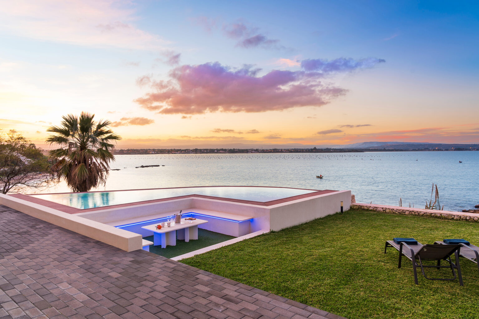 Stunning waterfront view featuring a modern infinity pool, a cozy outdoor seating area with a table and drinks, and a lush green lawn under a colorful sunset sky. | Only on SopranoVillas