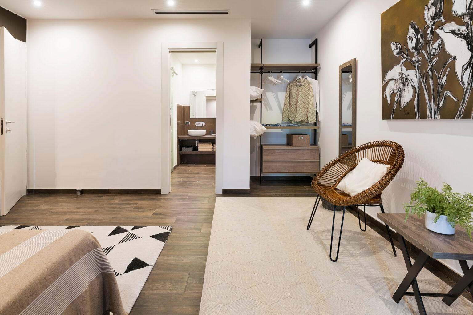Modern bedroom interior featuring a cozy atmosphere, with a stylish bed, a wicker chair, and an artistic wall painting. The space includes wooden flooring, a soft area rug, and a practical closet area, creating a warm and inviting environment. | Only on SopranoVillas