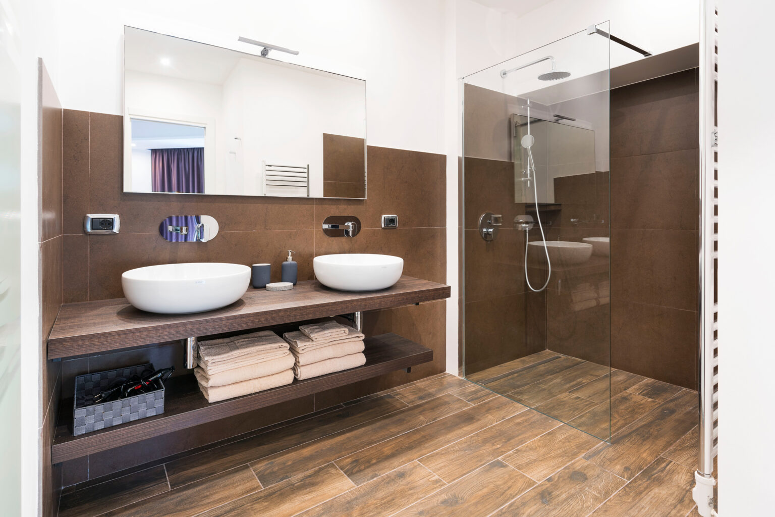 Modern bathroom interior featuring a double sink with round basins, a large mirror, and a glass shower enclosure. The space is designed with brown tiled walls, wooden flooring, and neatly stacked towels on a wooden shelf. | Only on SopranoVillas