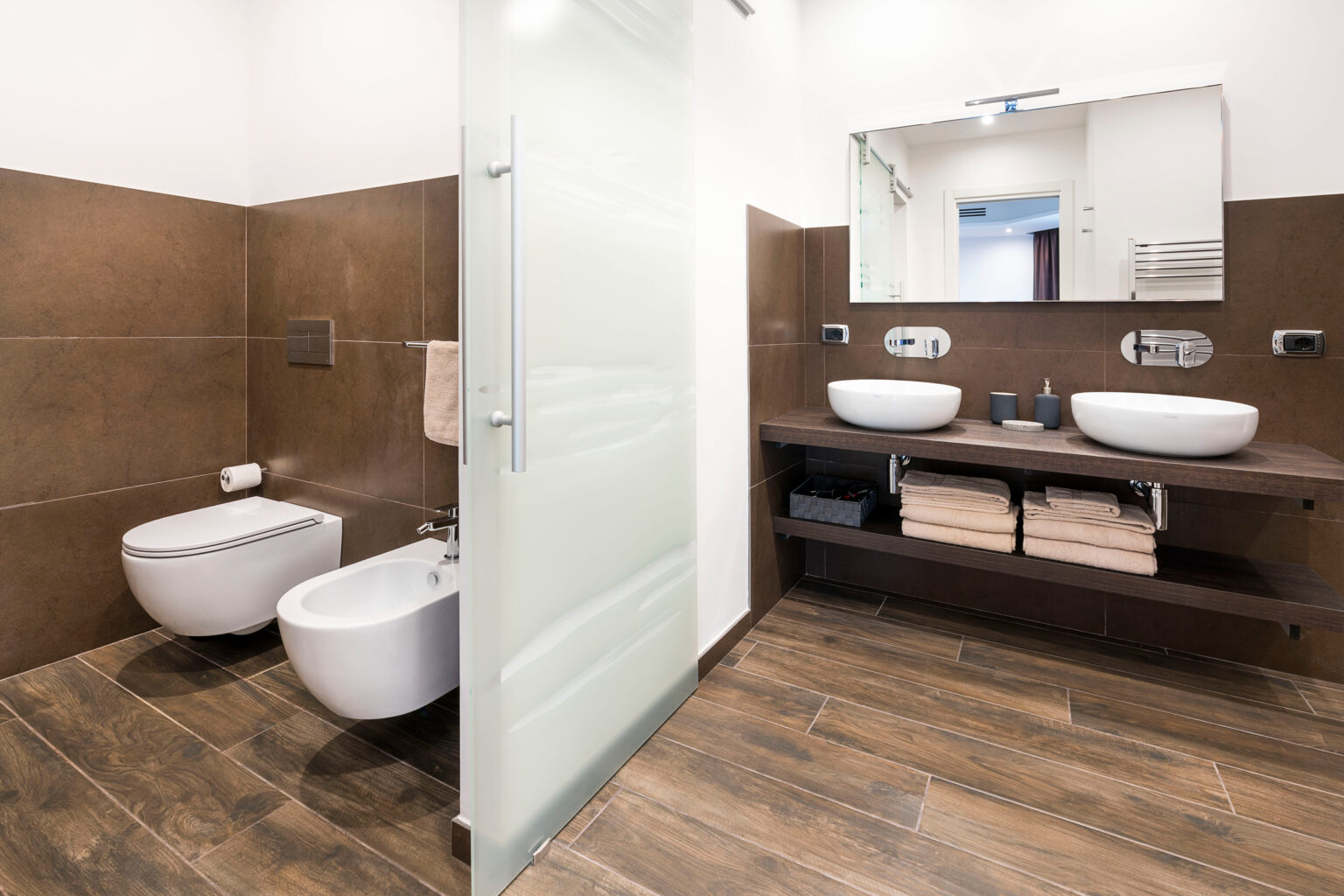 Modern bathroom design featuring a glass door, wall-mounted bidet, and two vessel sinks on a wooden vanity. The space is accented with brown tiled walls and wooden flooring, creating a stylish and contemporary aesthetic. | Only on SopranoVillas