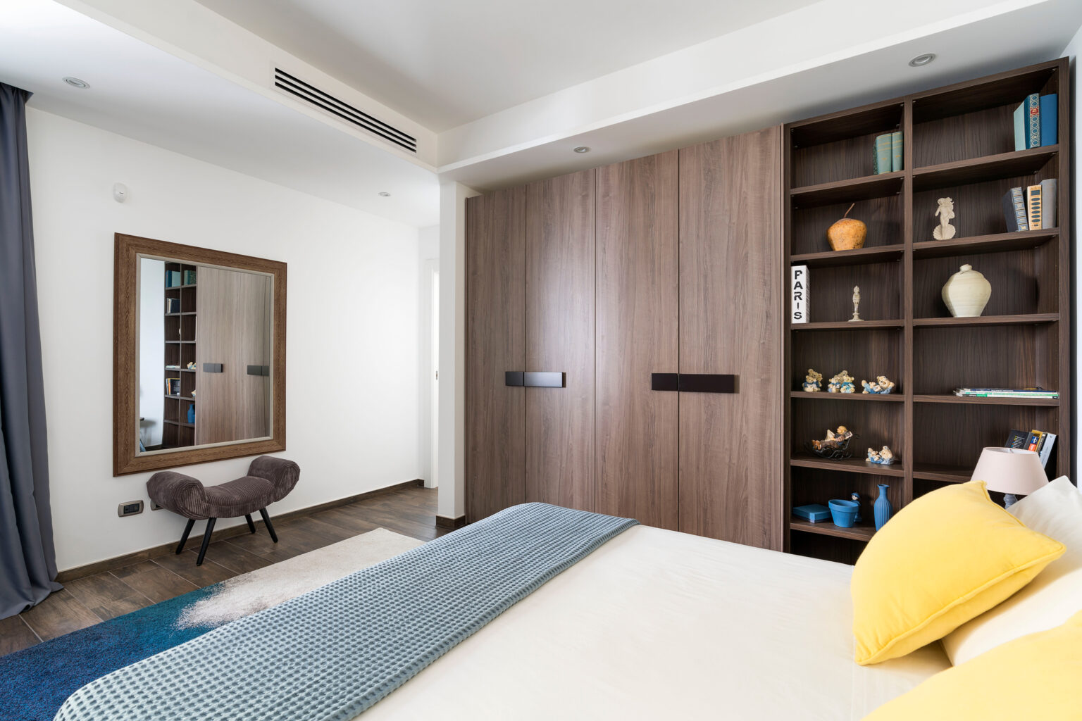 Modern bedroom interior featuring a wooden wardrobe, a decorative bookcase with assorted items, and a cozy bench. The room is adorned with a large mirror and a blue textured throw on the bed, complemented by yellow pillows. Natural light enhances the contemporary design and warm color palette. | Only on SopranoVillas
