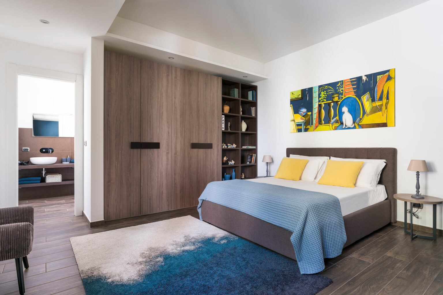 Modern bedroom featuring a queen-sized bed with yellow pillows, a cozy blue throw blanket, and a stylish area rug. The room includes a large wooden wardrobe, a bookshelf with decorative items, and a vibrant artwork on the wall. Natural light enhances the contemporary design and inviting atmosphere. | Only on SopranoVillas