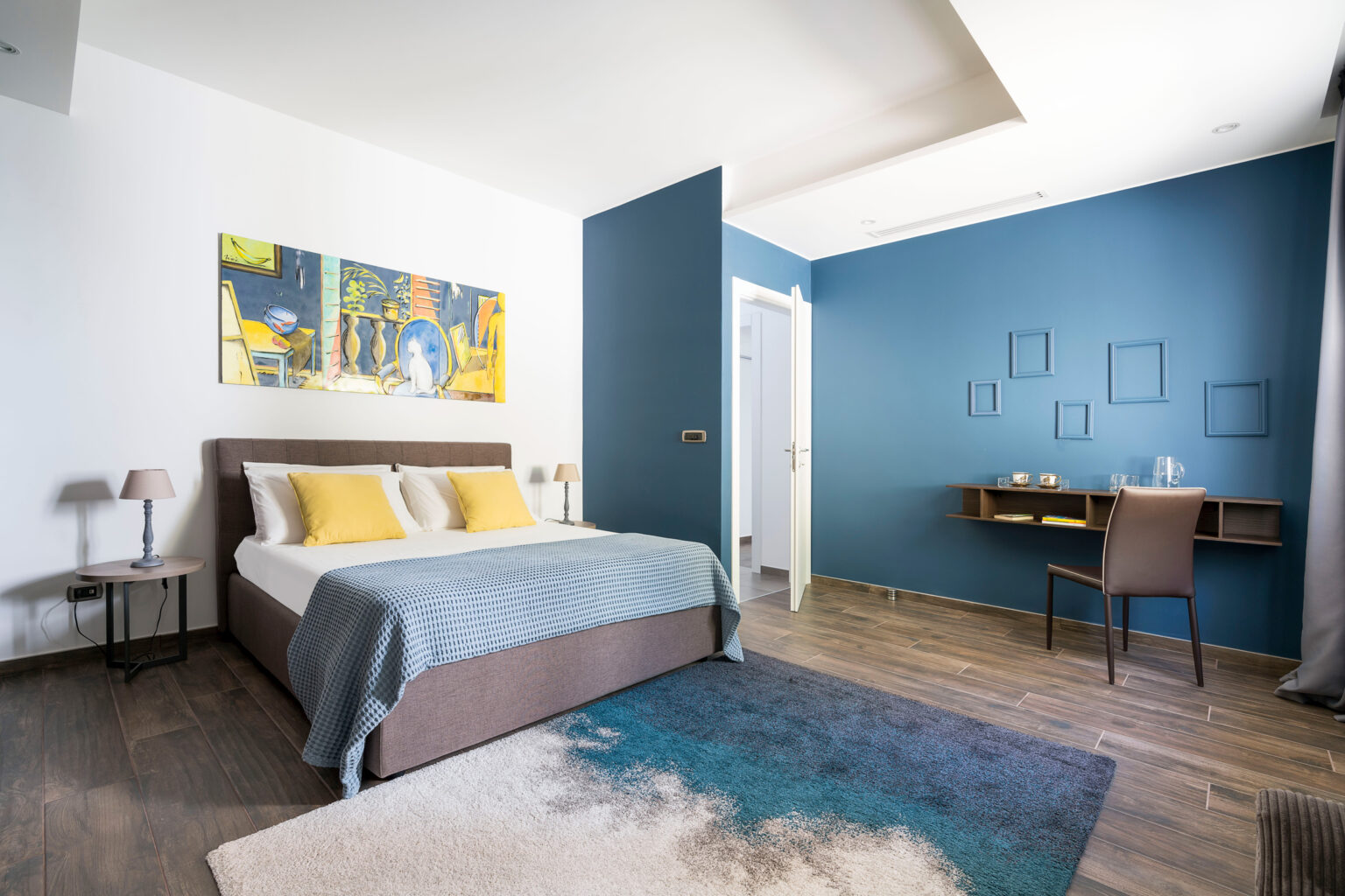 Modern hotel room featuring a queen-sized bed with yellow accent pillows, a blue throw blanket, and stylish furnishings. The room includes a decorative painting on the wall, a small bedside table with a lamp, and a workspace with a chair and shelves. The color scheme combines light and dark blue walls with wooden flooring and a plush area rug, creating a cozy atmosphere. | Only on SopranoVillas