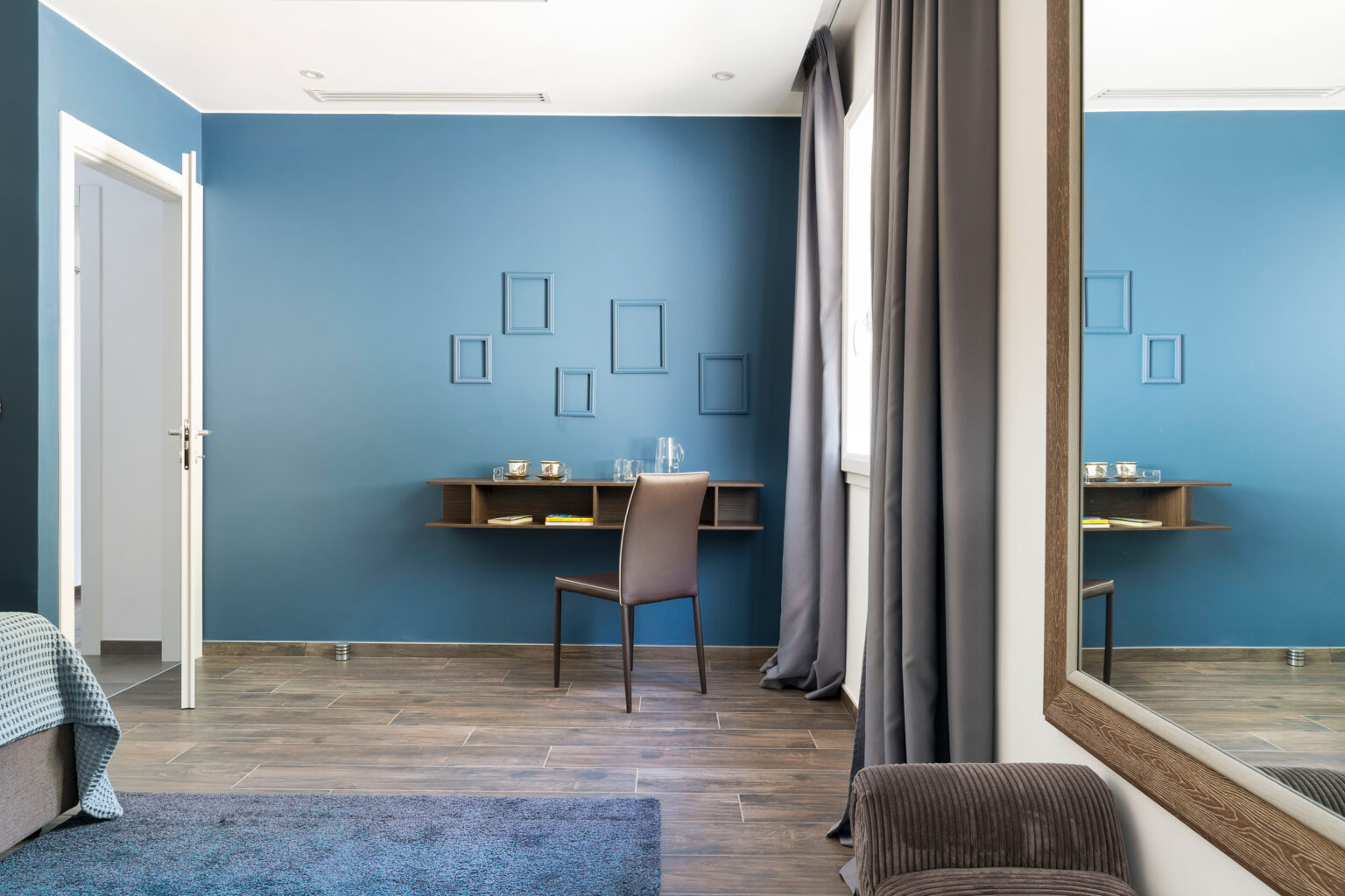 Modern bedroom interior featuring a blue accent wall, a wooden desk with decorative items, a comfortable chair, and a large mirror reflecting the space. The room has a cozy atmosphere with natural light and stylish decor elements. | Only on SopranoVillas
