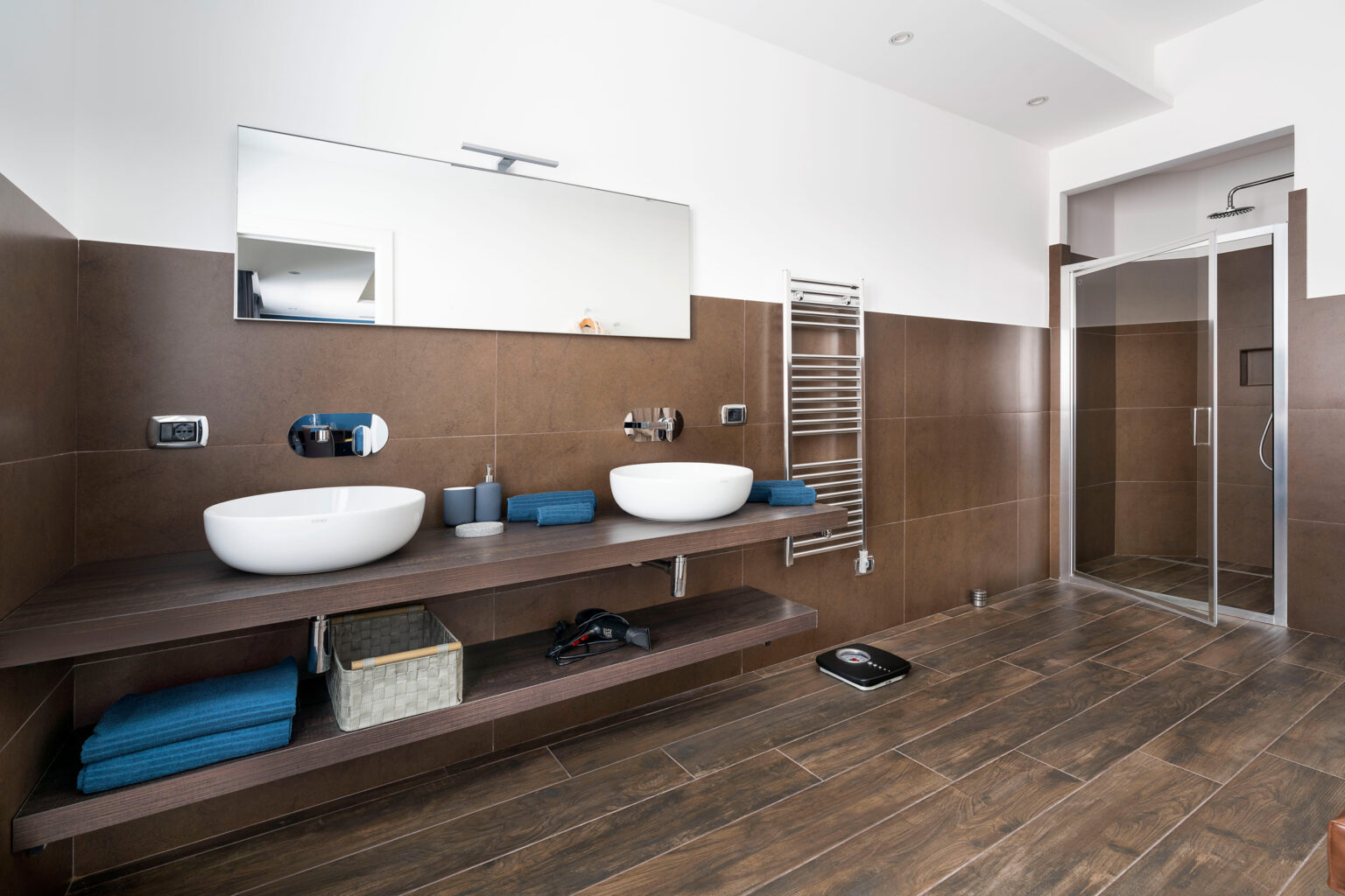 Modern bathroom design featuring dual vessel sinks on a wooden countertop, a large mirror, stylish towel racks, and a glass shower enclosure. The space is accented with blue towels and a wooden floor, creating a contemporary and inviting atmosphere. | Only on SopranoVillas
