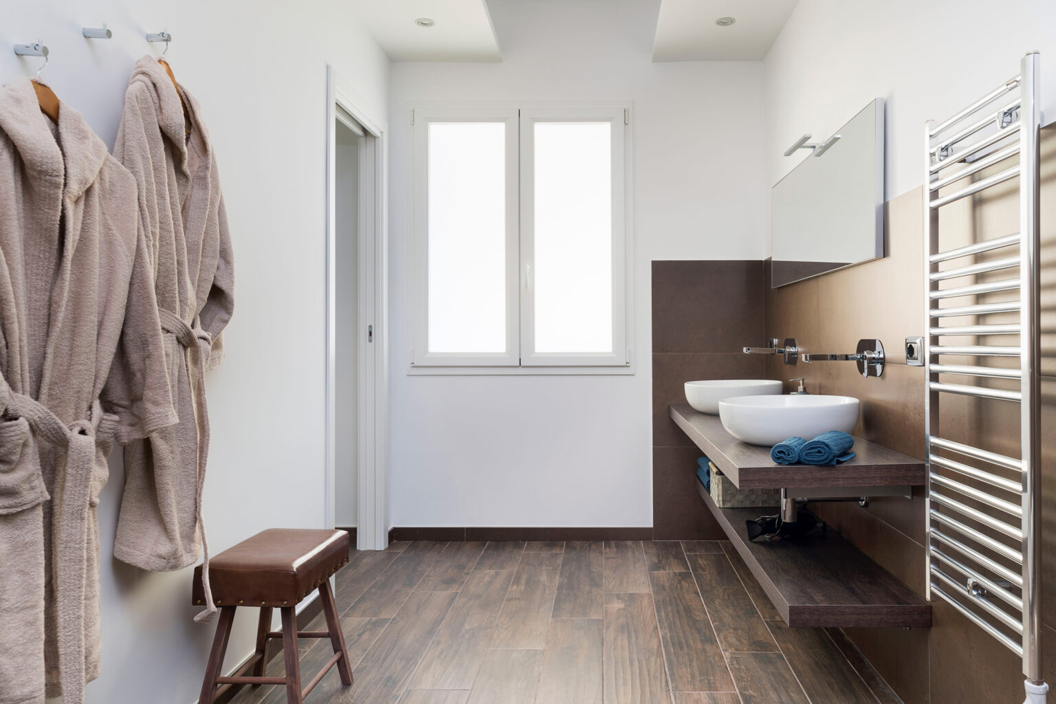 Modern bathroom interior featuring wooden flooring, dual sinks, a sleek mirror, and a heated towel rack. Beige bathrobes hang on hooks, and large windows provide natural light. | Only on SopranoVillas