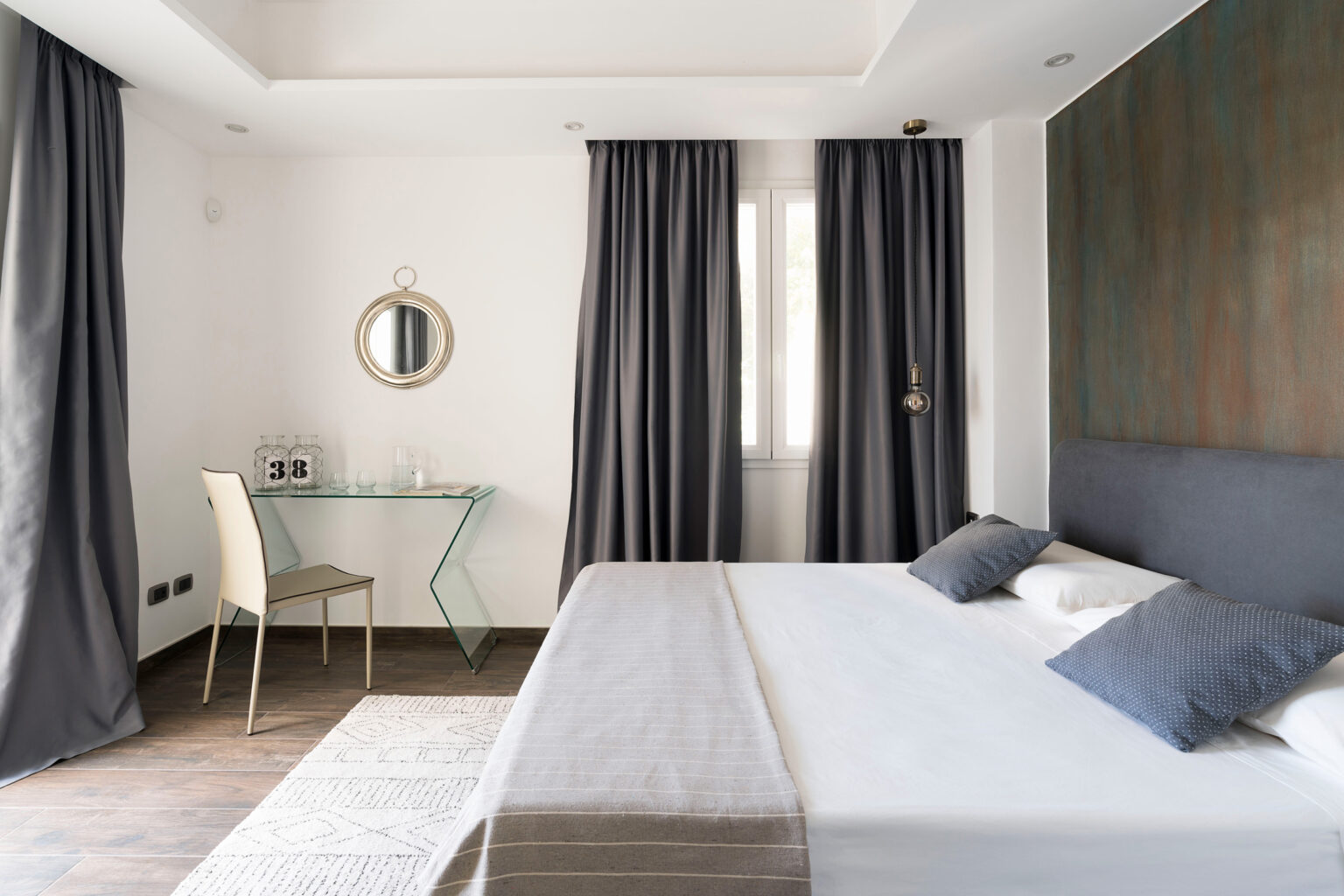 Modern bedroom interior featuring a queen-sized bed with decorative pillows, a stylish glass desk with a chair, and elegant gray curtains. The room is well-lit with natural light from a window, complemented by a contemporary mirror and a unique pendant light fixture, creating a serene and inviting atmosphere. | Only on SopranoVillas