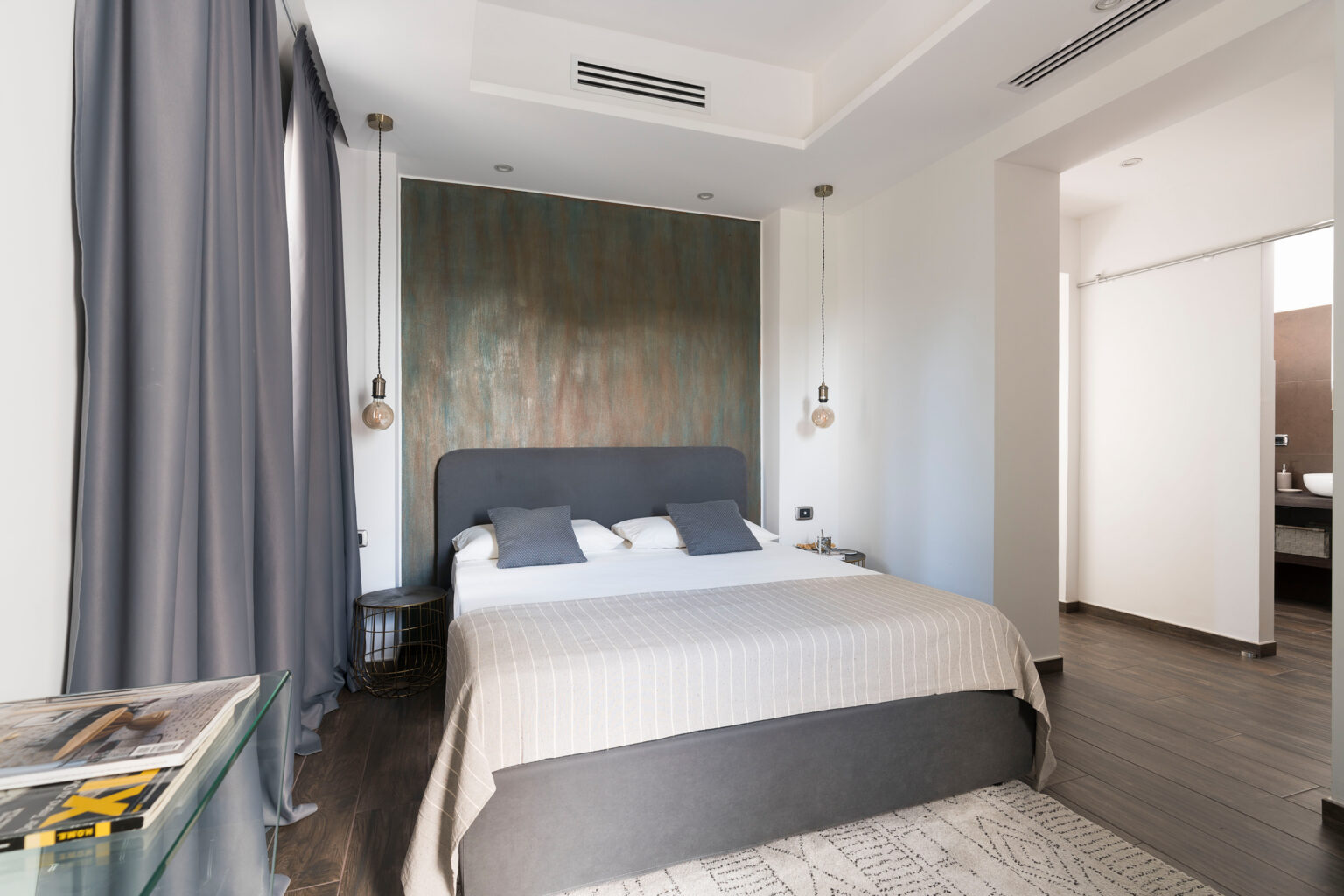 Modern bedroom interior featuring a gray bed with decorative pillows, a textured accent wall, stylish pendant lighting, and elegant curtains. A glass side table displays magazines, while the wooden floor adds warmth to the space. | Only on SopranoVillas