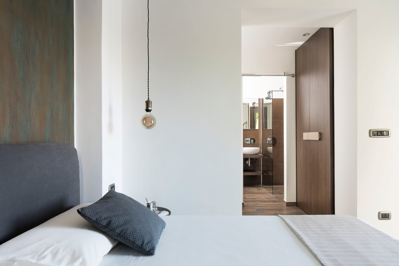 Modern bedroom interior featuring a minimalist design with a gray bed, decorative pillow, and a pendant light, leading to a bathroom visible through an open door. | Only on SopranoVillas