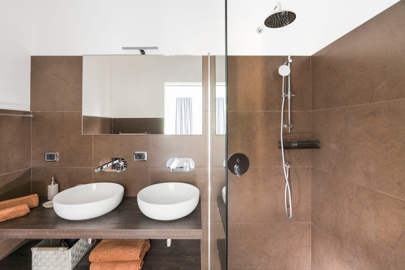 Modern bathroom design featuring dual white vessel sinks, a large mirror, and a glass shower enclosure. The space is accented with brown tiled walls and wooden shelving, complemented by neatly arranged orange towels. Ideal for showcasing contemporary interior aesthetics. | Only on SopranoVillas