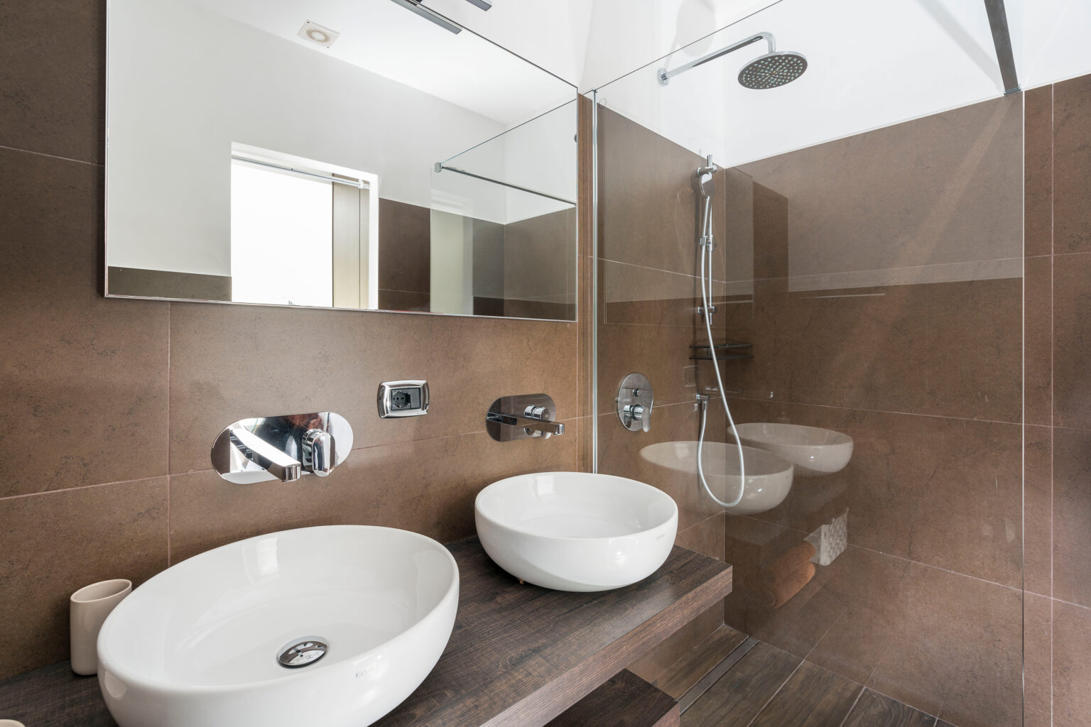 Modern bathroom design featuring a sleek layout with two white vessel sinks on a wooden countertop, a glass shower with a rainfall showerhead, and stylish chrome fixtures against a warm brown tiled backdrop. Natural light enters through a window, enhancing the contemporary aesthetic. | Only on SopranoVillas