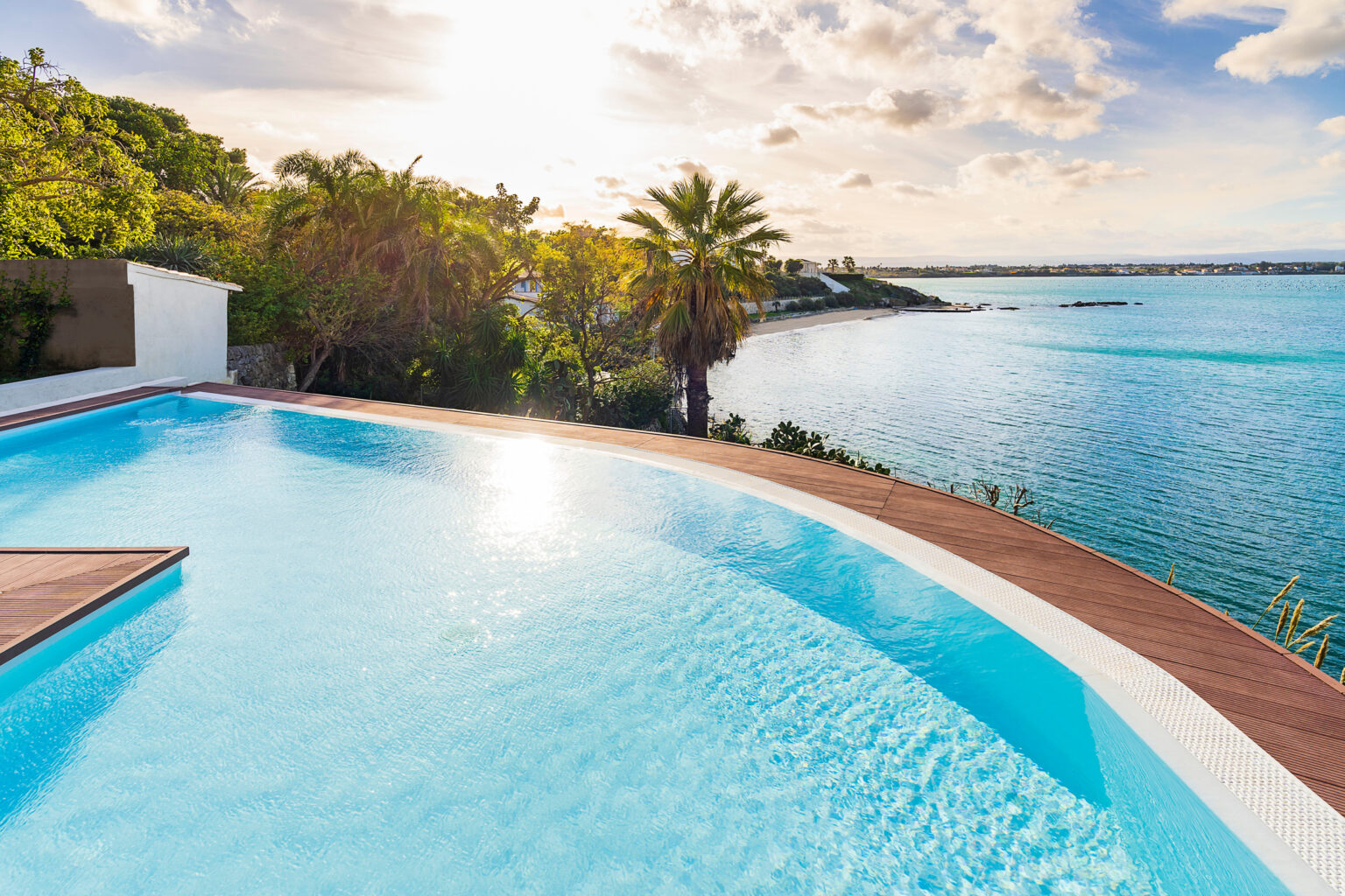 Scenic view of a luxury infinity pool overlooking a tranquil ocean, surrounded by lush greenery and palm trees, with a sunset sky in the background. Ideal setting for a relaxing getaway or vacation rental. | Only on SopranoVillas