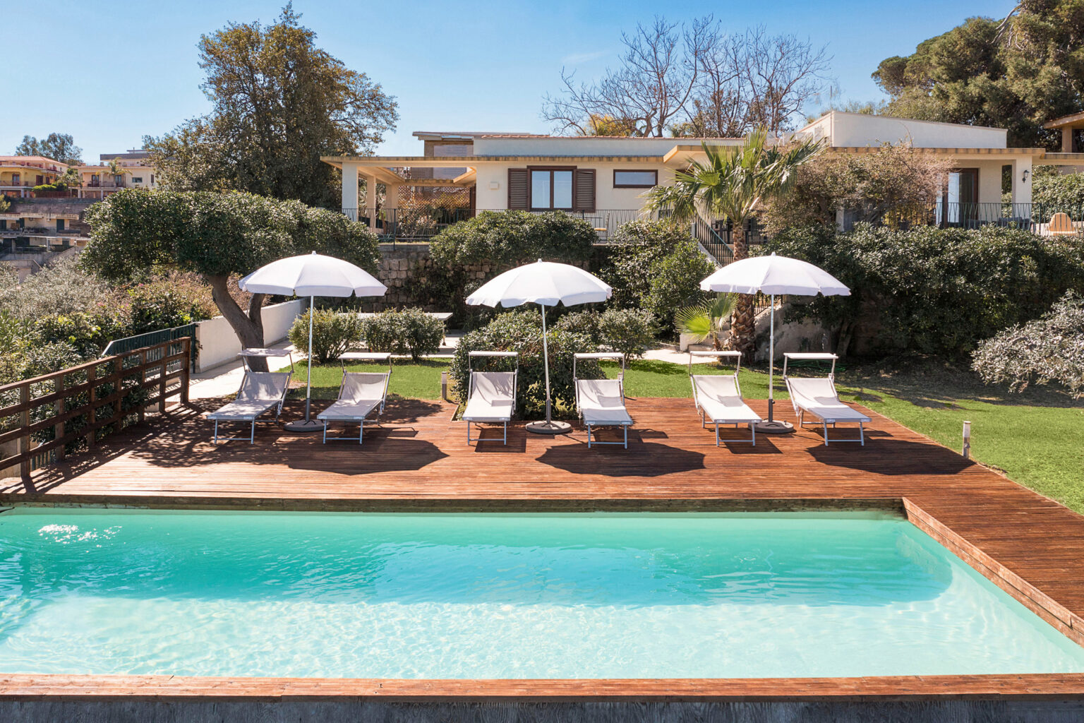 Luxury villa with a swimming pool, wooden deck, and sun loungers under white umbrellas, surrounded by lush greenery and a clear blue sky. Perfect for relaxation and outdoor gatherings. | Only on SopranoVillas