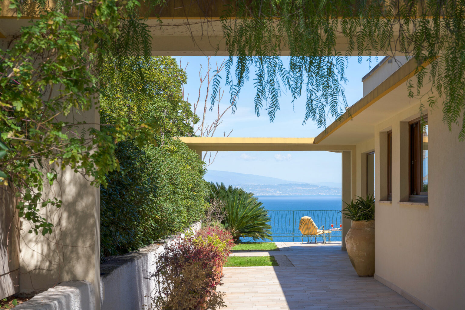 Modern coastal home exterior with a pathway leading to a stunning ocean view, framed by lush greenery and contemporary architectural elements. | Only on SopranoVillas