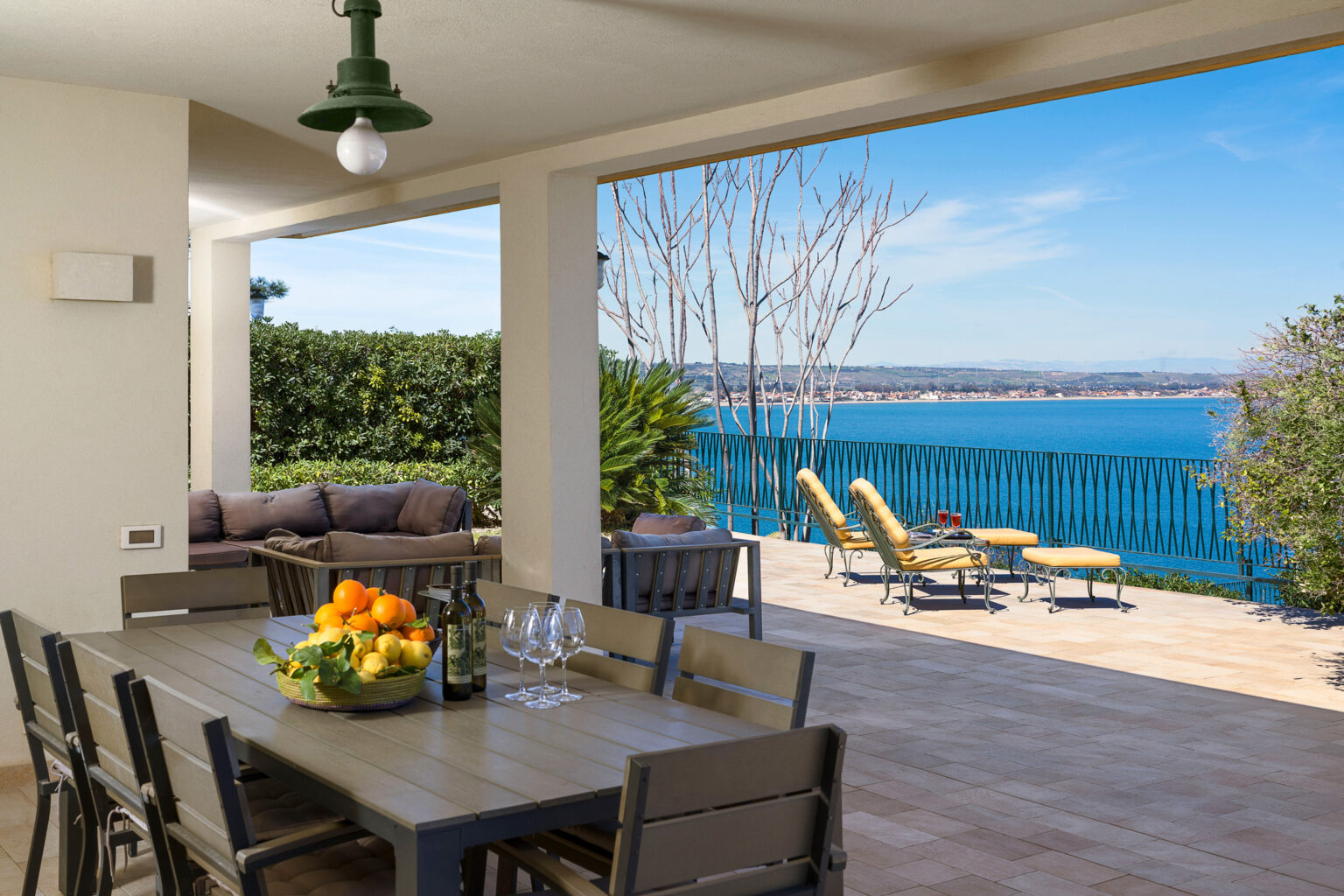 Spacious outdoor dining area with a wooden table set for a meal, overlooking a serene lake view, featuring comfortable seating and sun loungers, ideal for relaxation and entertaining. | Only on SopranoVillas