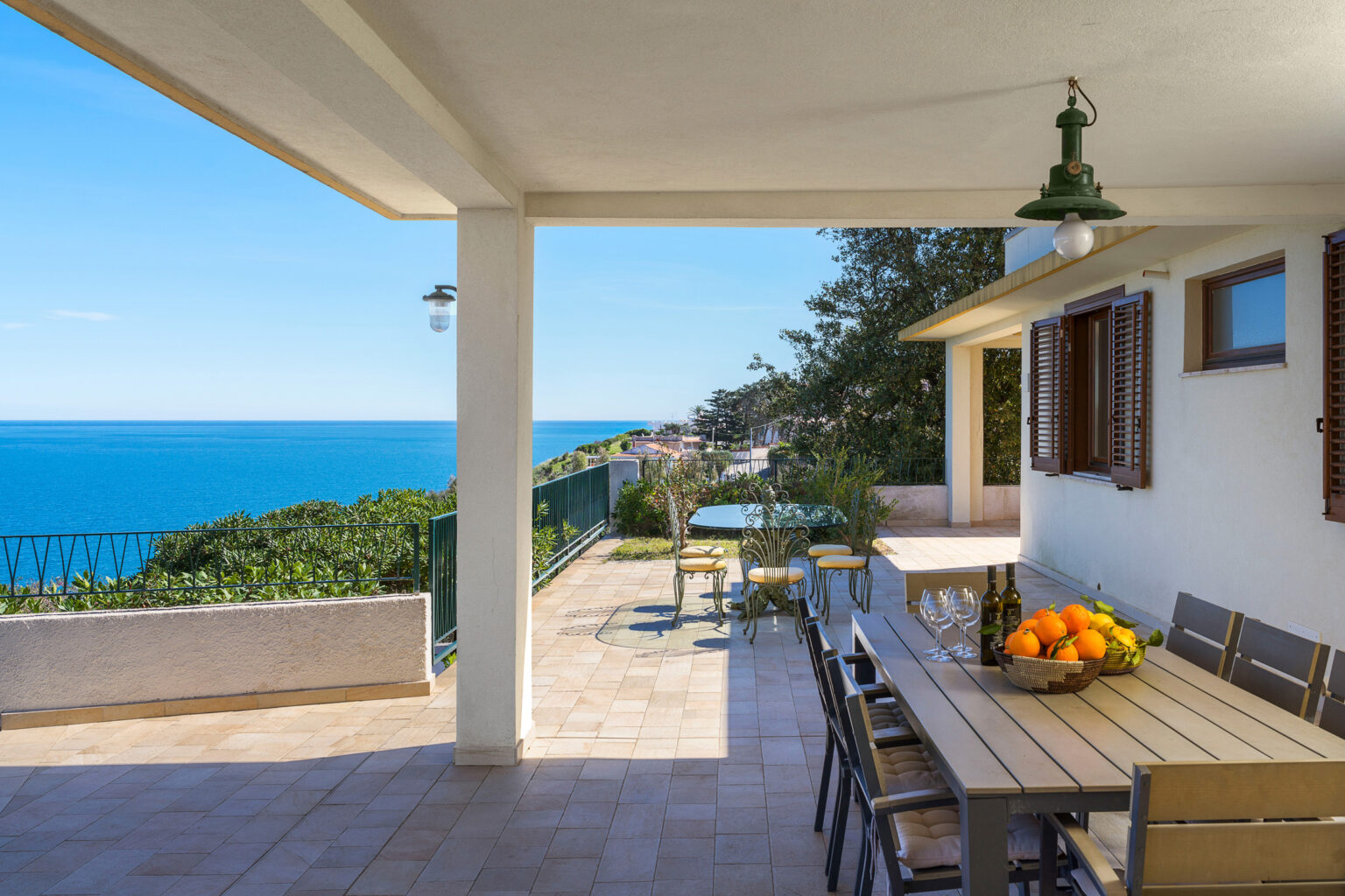 Cozy outdoor terrace with a dining table and fruit basket, overlooking a stunning ocean view and lush greenery, perfect for relaxation and entertaining. | Only on SopranoVillas