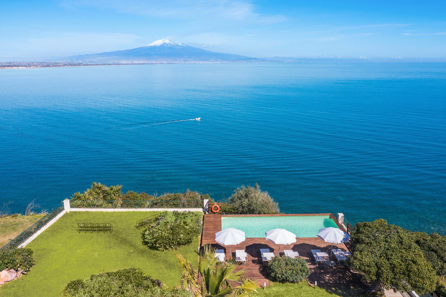 Aerial view of a luxurious villa with a swimming pool overlooking the blue sea and Mount Etna in the background, featuring sun loungers and umbrellas on a wooden deck surrounded by greenery. | Only on SopranoVillas