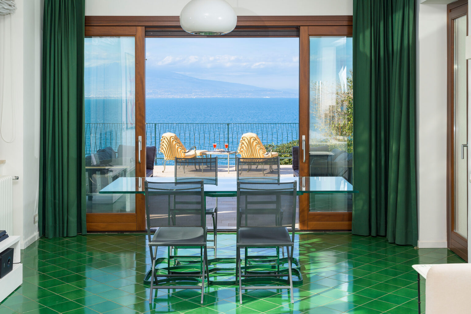 Bright and modern dining area featuring a glass table and four black chairs, with large glass doors leading to a balcony overlooking the sea. The interior is adorned with green curtains and glossy green tiled flooring, creating a vibrant atmosphere complemented by a stunning ocean view. | Only on SopranoVillas