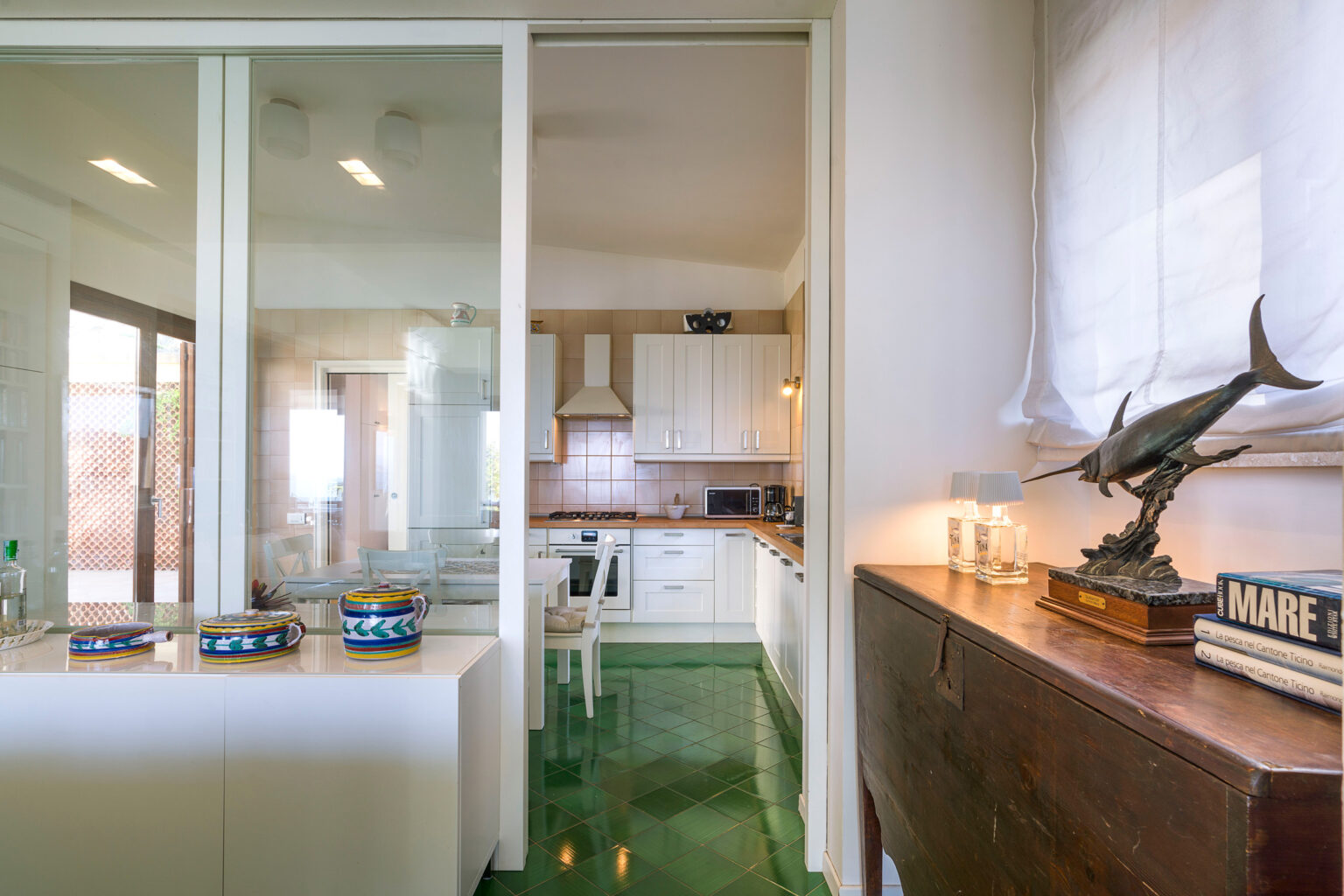 Modern kitchen interior featuring white cabinetry, a green floor, and a decorative fish sculpture on a wooden side table. Natural light streams in through large windows, creating an inviting atmosphere. | Only on SopranoVillas