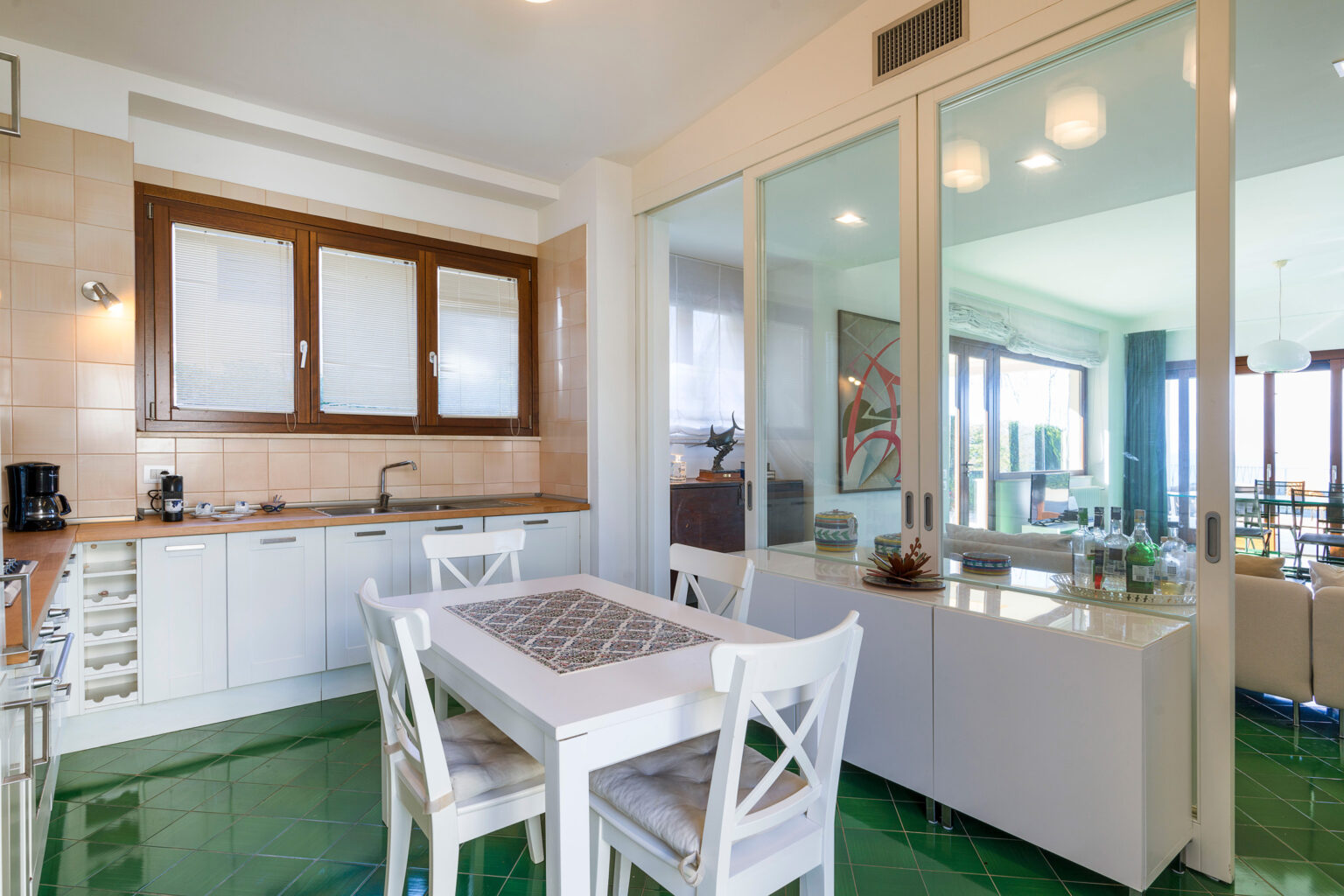 Modern kitchen with white cabinetry, a wooden dining table, and green flooring, featuring large windows that provide natural light and a view of the living area. | Only on SopranoVillas